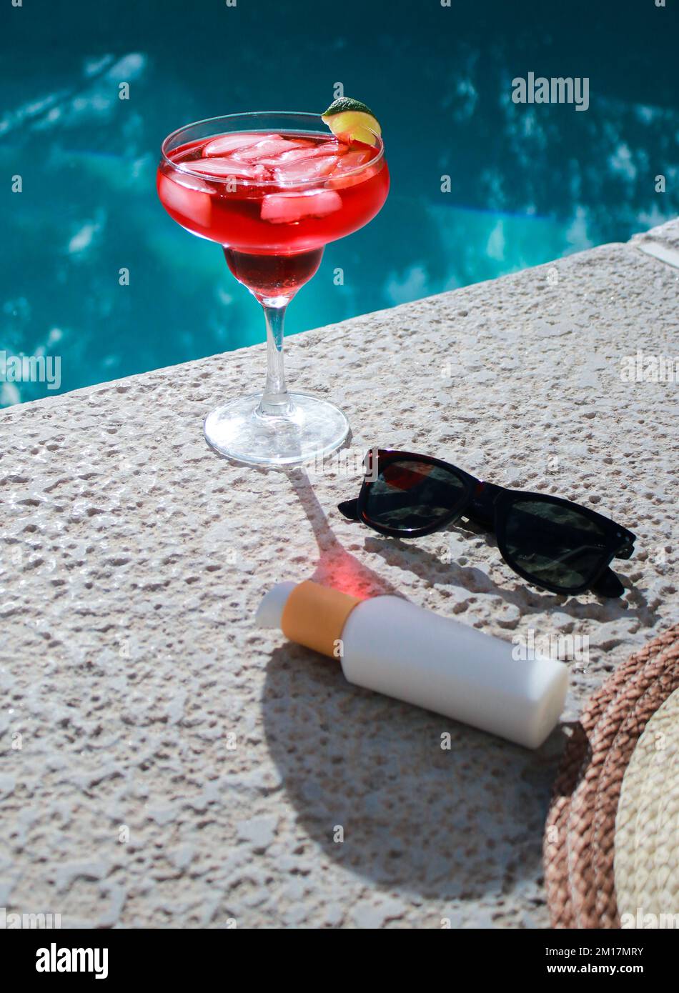 Rinfrescante cocktail rosso in piscina con crema solare, occhiali da sole e cappello di paglia Foto Stock