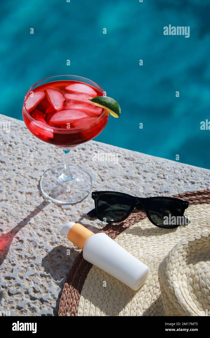 Rinfrescante cocktail rosso in piscina con crema solare, occhiali da sole e cappello di paglia Foto Stock
