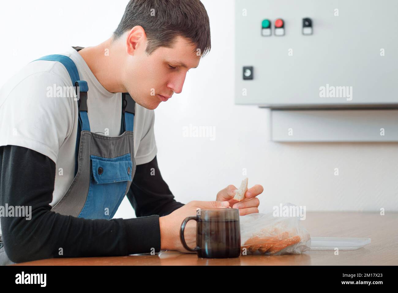 Pausa pranzo. Mangiare sul posto di lavoro durante il lavoro. L'uomo caucasico in tuta da lavoro siede al tavolo nella sala di produzione e mangia dal contenitore. Il lavoratore ha pranzo in sala pausa. Foto Stock