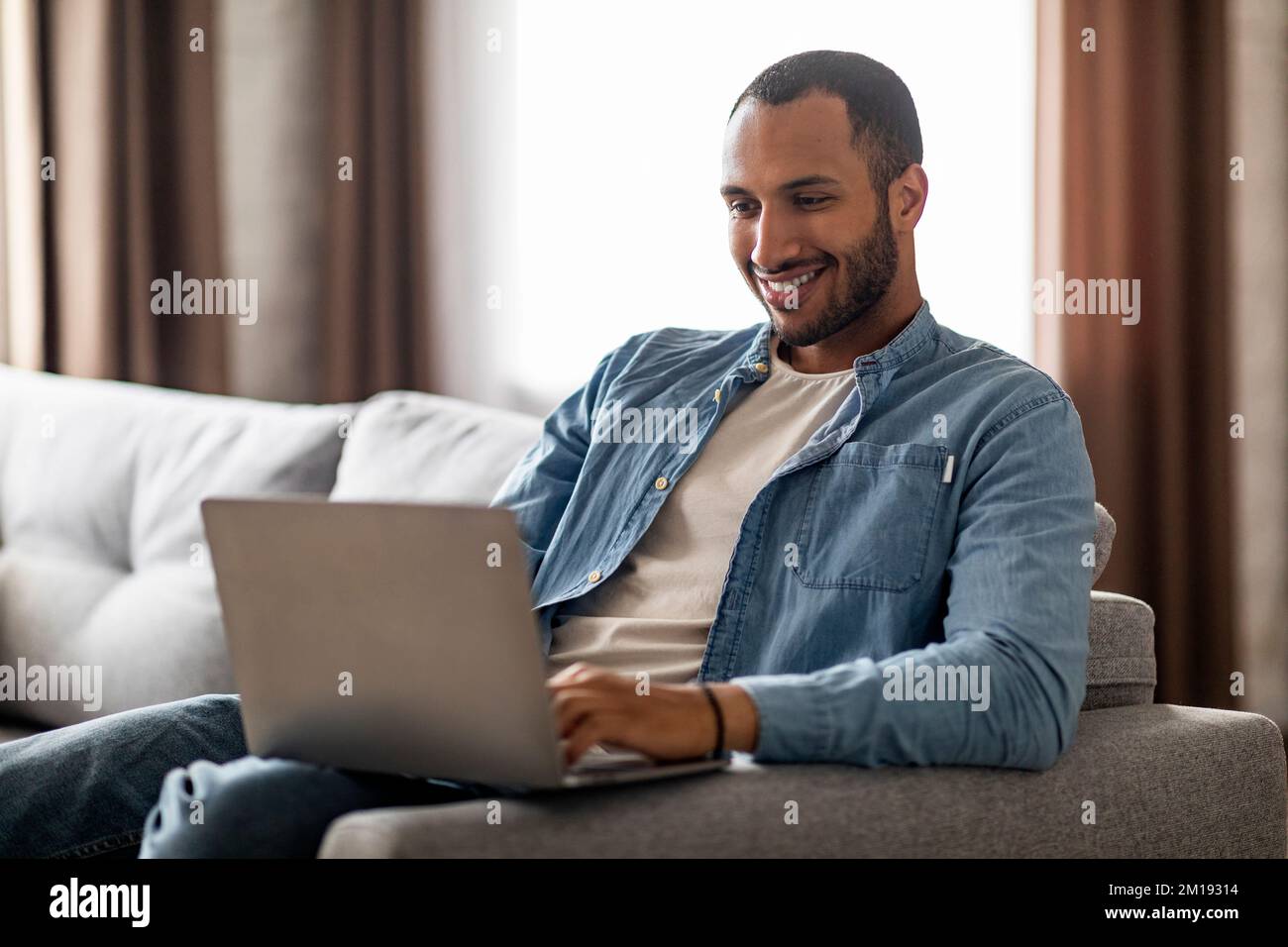 Corrispondenza online. Sorridente uomo nero seduto sul divano e digitando sul computer portatile Foto Stock