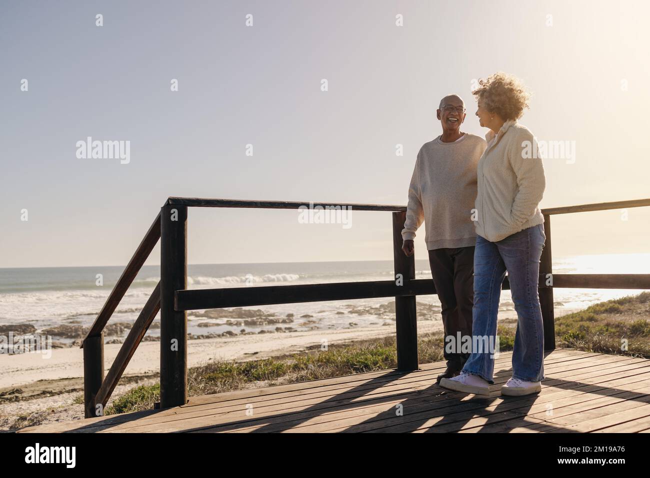 Coppia matura sorridente mentre si fa una passeggiata lungo un ponte pedonale in legno sulla spiaggia. Coppia anziana romantica godendo di qualità di spesa t Foto Stock