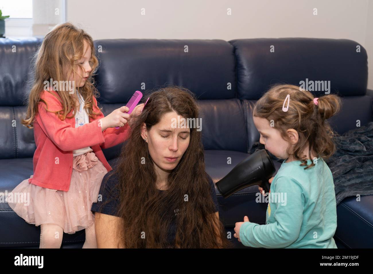 Una madre e le sue due figlie giocano e interagiscono insieme, con una ragazza che pettina i capelli bagnati e l'altra che asciuga i capelli con un asciugacapelli Foto Stock
