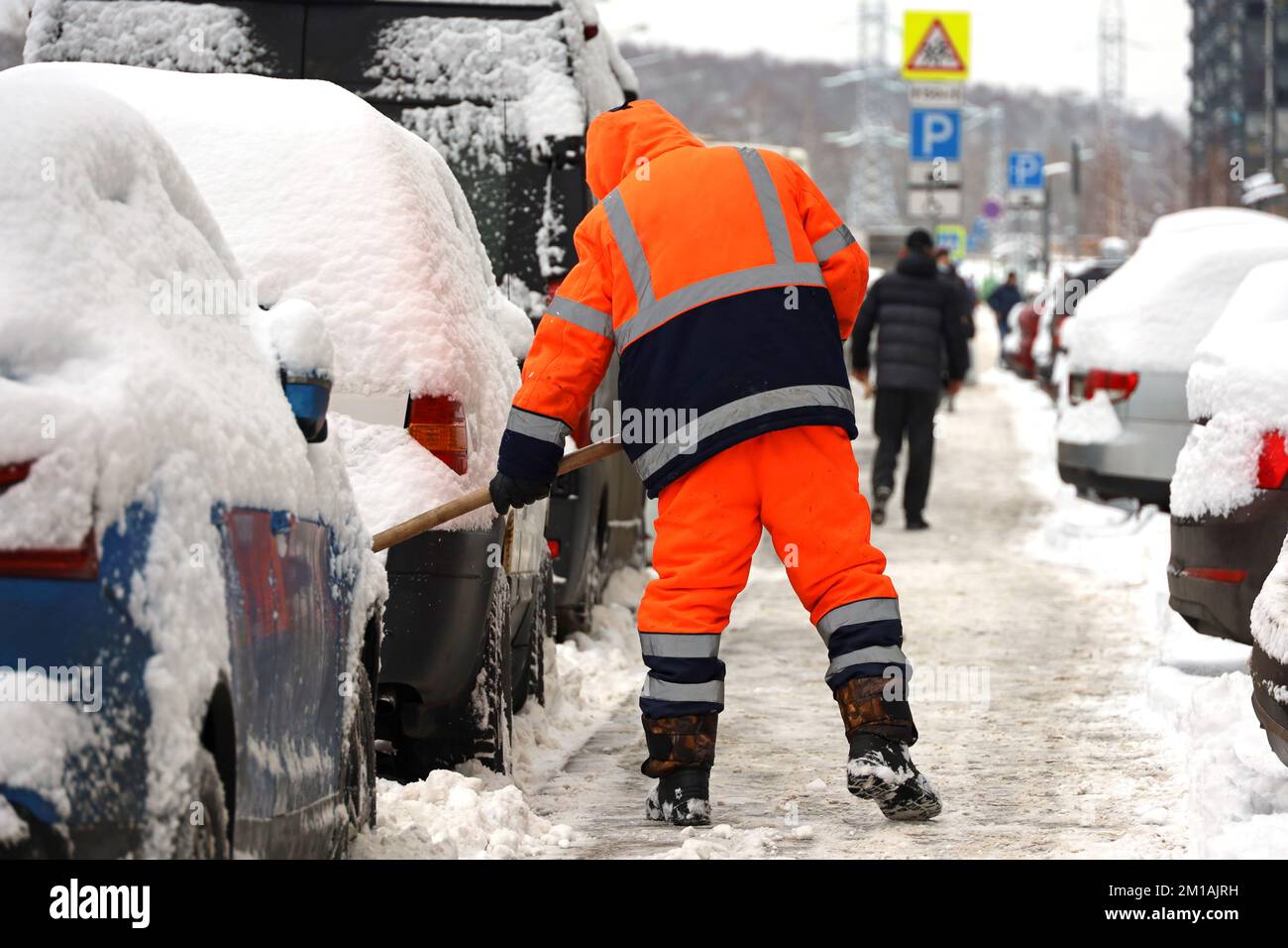 Uomo operaio in uniforme arancione con una pala che pulisce la strada vicino al parcheggio auto. Rimozione della neve nella città invernale durante la tempesta di neve Foto Stock
