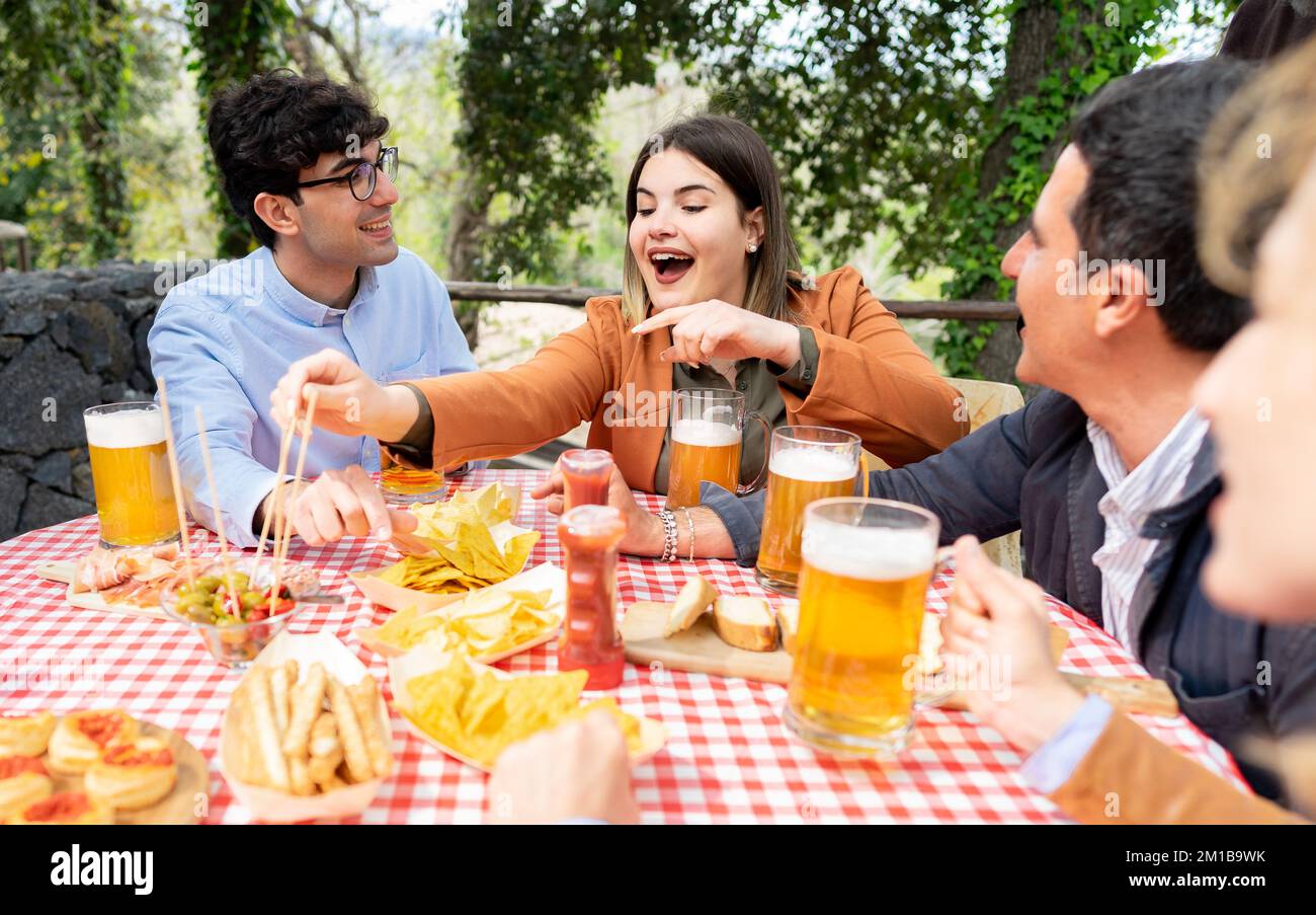 Gruppo di amici di età mista bere pinte di birra al pub fuori lato mentre mangiare tapas- felice gente divertirsi insieme nel giardino della fattoria Foto Stock