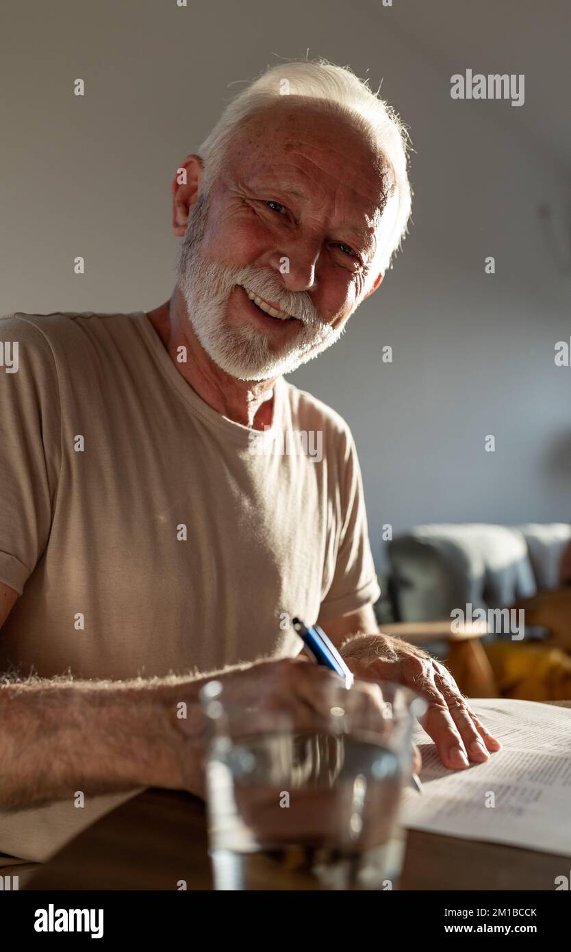 Uomo anziano soddisfatto seduto al tavolo a casa e leggendo carta con sorriso sul viso Foto Stock