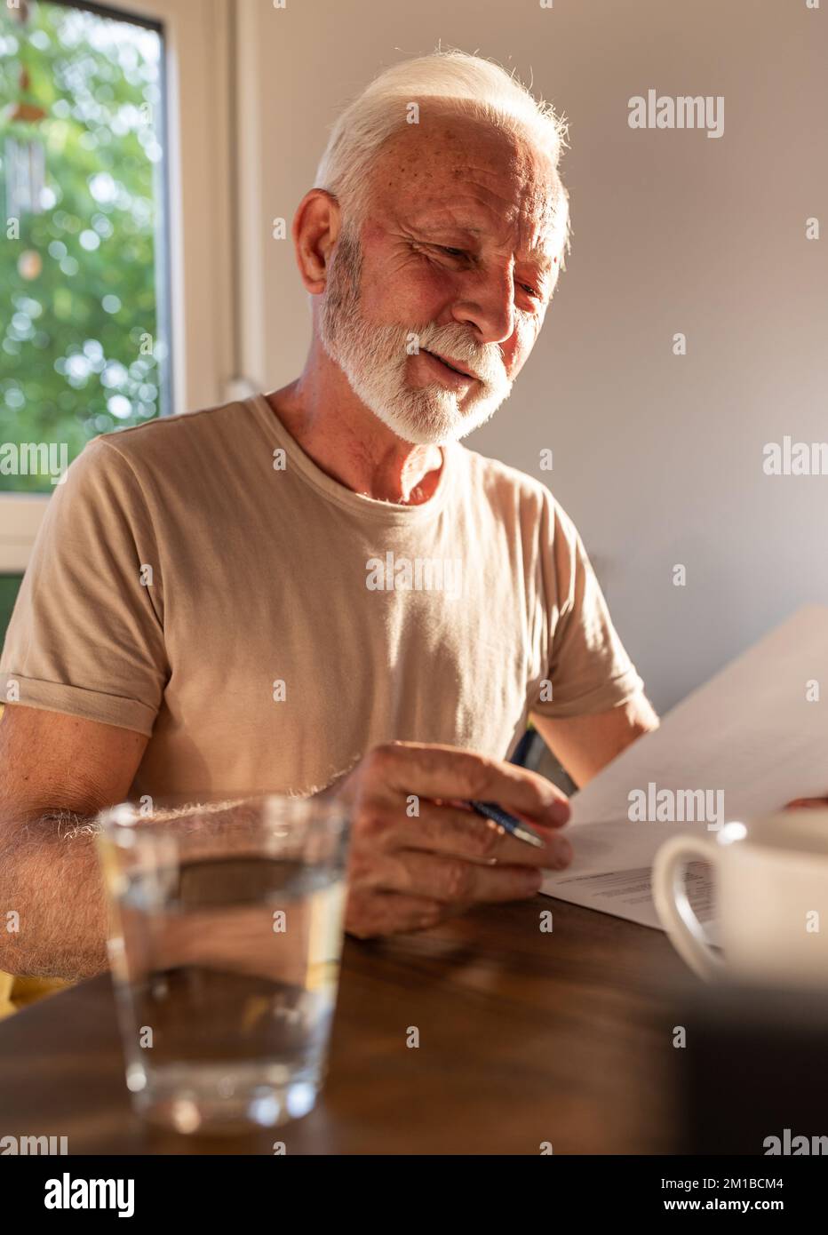 Uomo anziano soddisfatto seduto al tavolo a casa e leggendo carta con sorriso sul viso Foto Stock