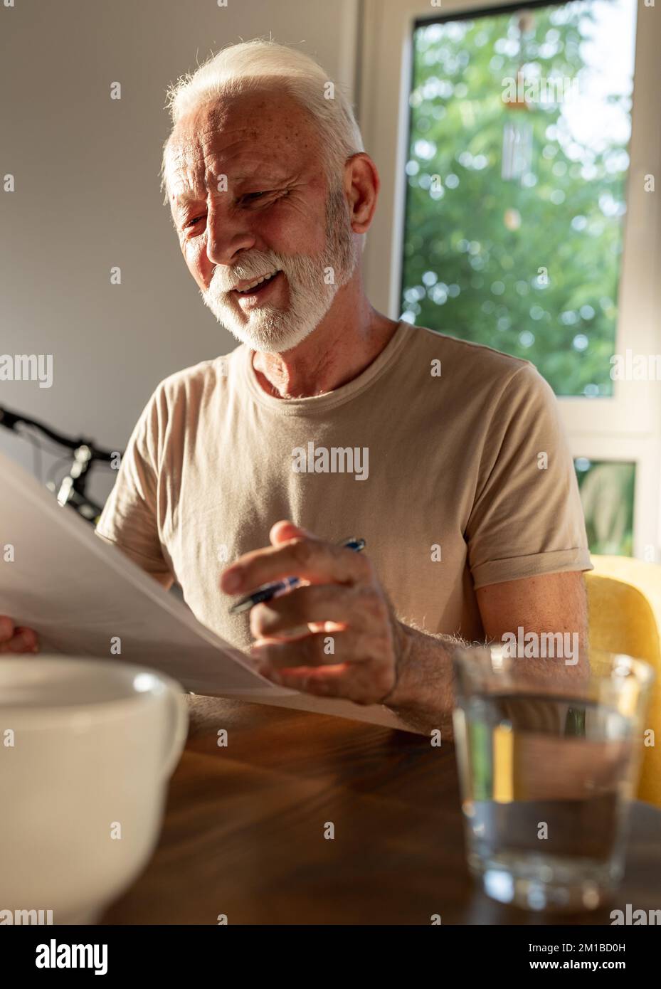 Uomo anziano soddisfatto seduto al tavolo a casa e leggendo carta con sorriso sul viso Foto Stock