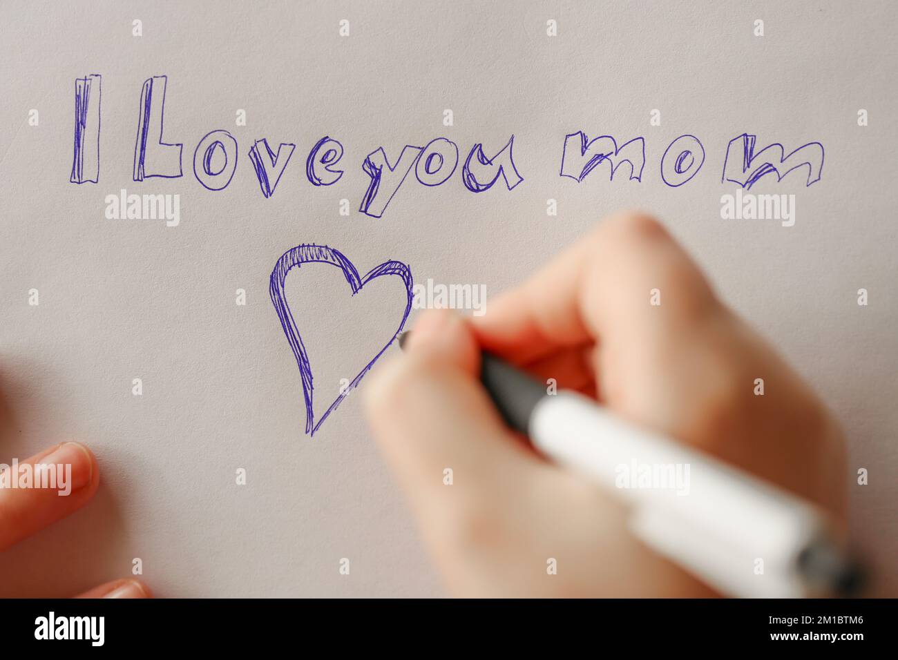 Madri day.moms day.i love you mom iscrizione su un foglio bianco e un cuore e una mano di bambini con una penna. Foto Stock