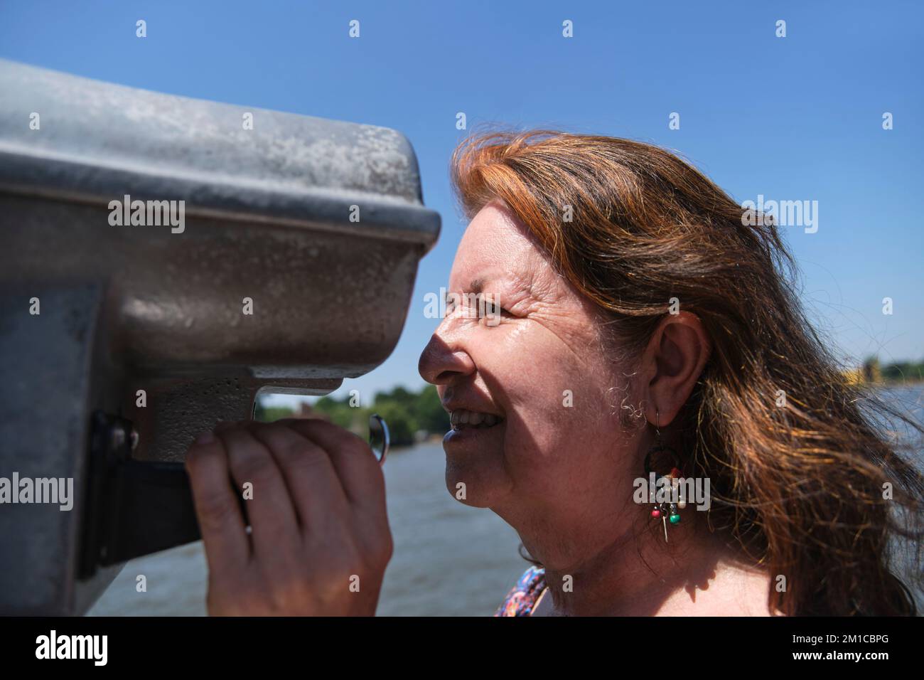 Ritratto di una donna matura ispanica che si diverte a guardare attraverso binocoli pubblici. Concetti di curiosità, osservazione, esplorazione, ricerca, viaggio. Foto Stock