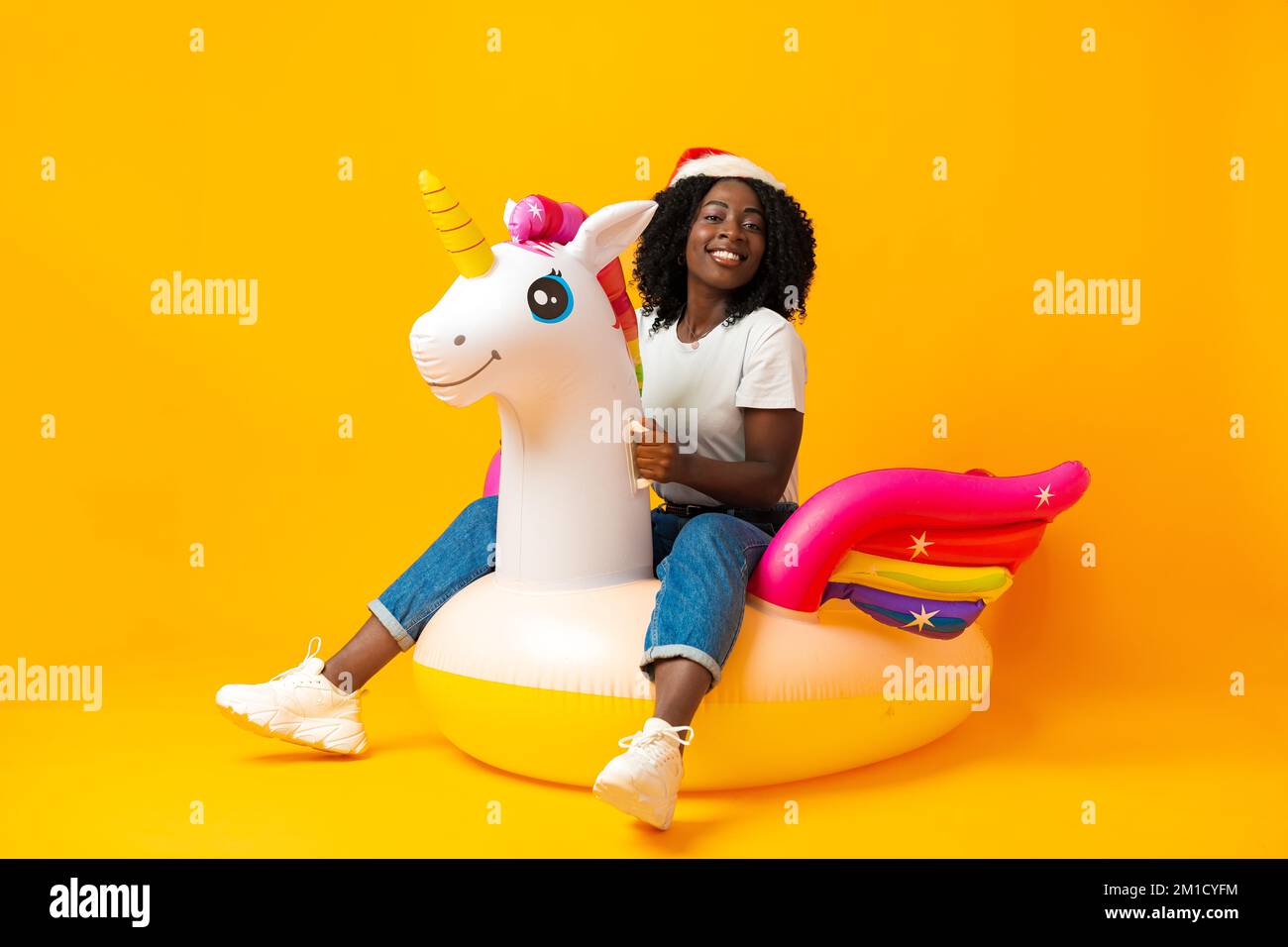 Donna africana seduta sul galleggiante dell'unicorno arcobaleno indossando il cappello di babbo natale in studio Foto Stock
