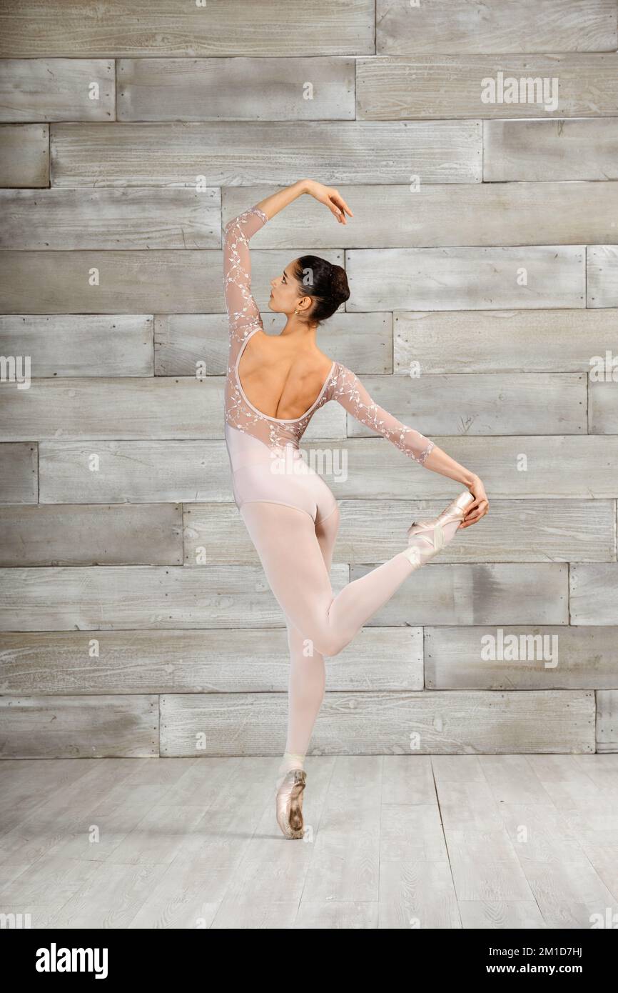 Corpo pieno giovane donna aggraziata che alza il braccio e la gamba mentre si posa sulla punta durante le prove di balletto in studio leggero Foto Stock