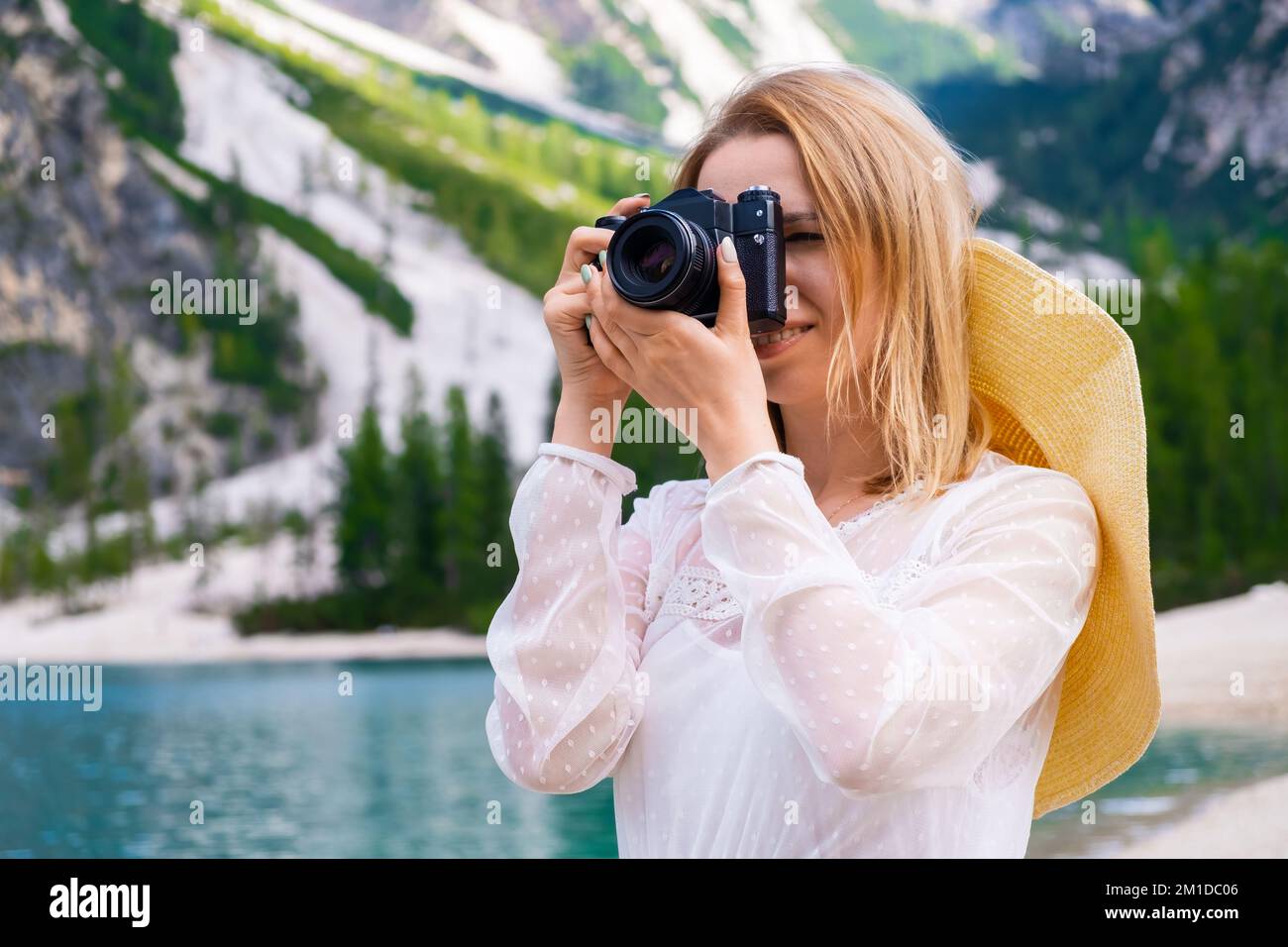 Sorridente bionda fotografa con una fotocamera professionale che scatta foto del lago. Ritratto di donna in cappello estivo e vestito bianco contro le Alpi Dolomiti. Foto Stock