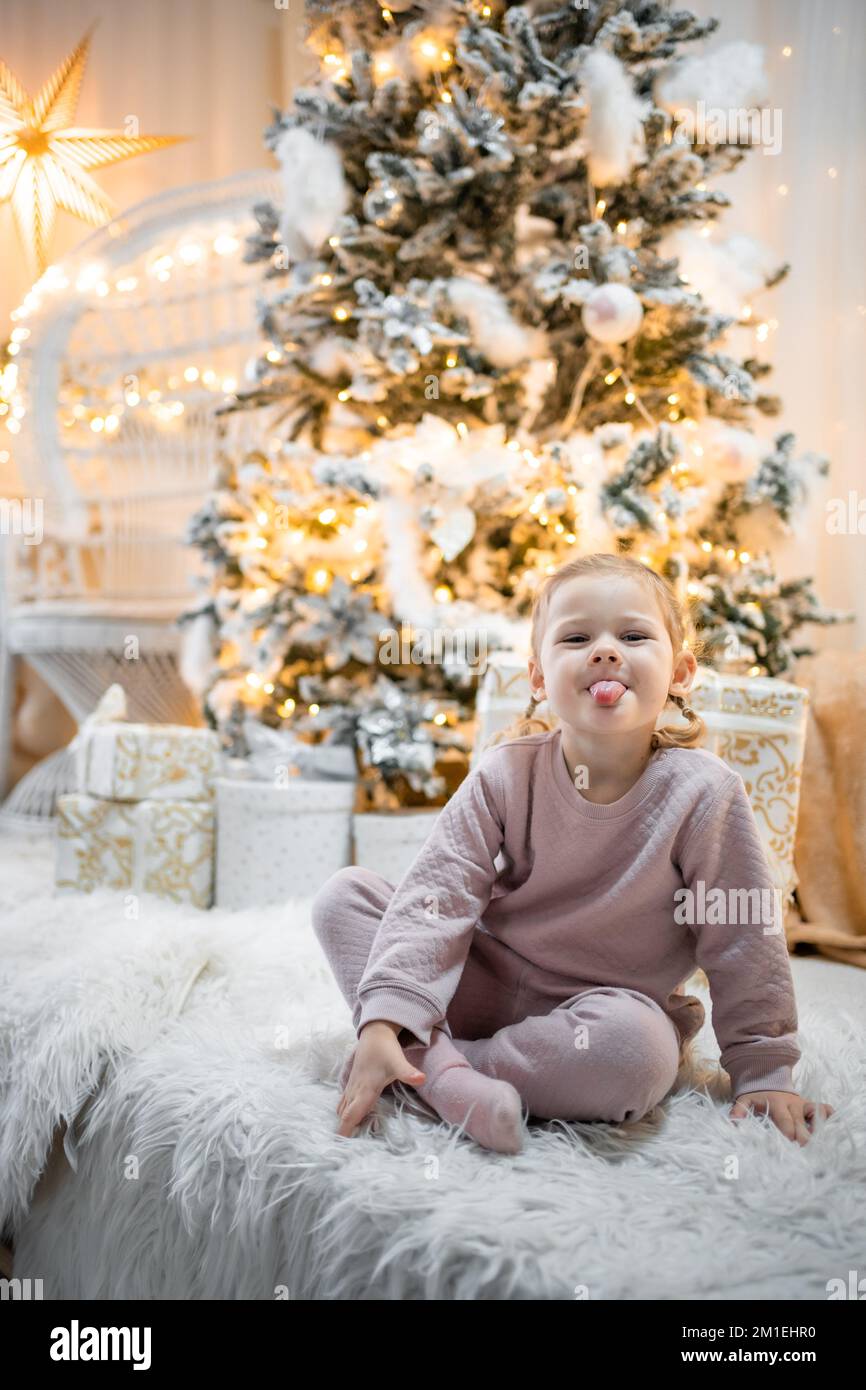 Molto bella ragazza piccola bionda affascinante in pigiama seduta sul letto di un bambino sullo sfondo di alberi di Natale in luminoso interno della casa Foto Stock
