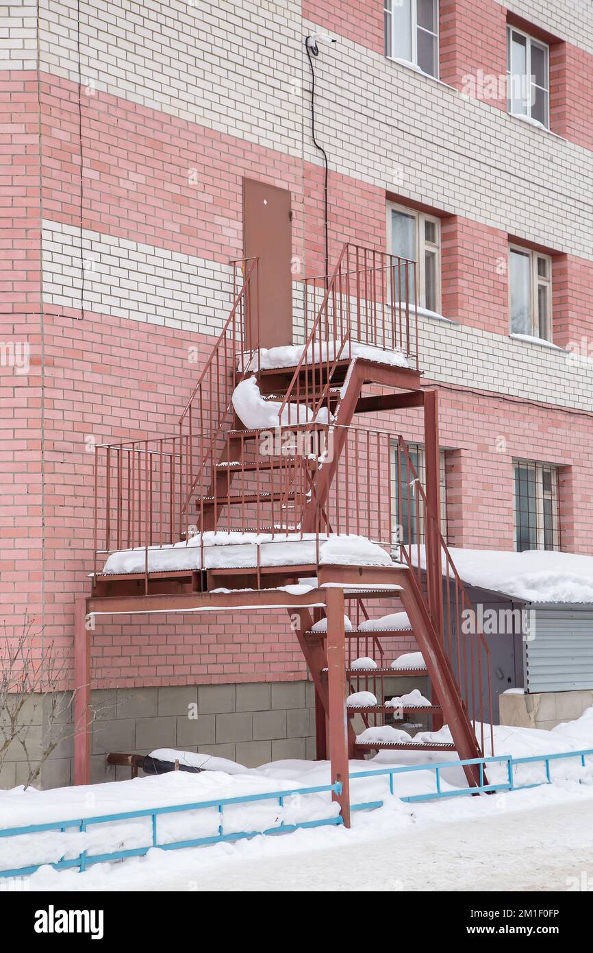 Scala in metallo di ricambio dall'esterno dell'edificio al secondo piano - foto in stock. Parete di un moderno edificio in mattoni con finestre di plastica. Inverno, neve tutto intorno. Luce calda e soffusa. Foto Stock