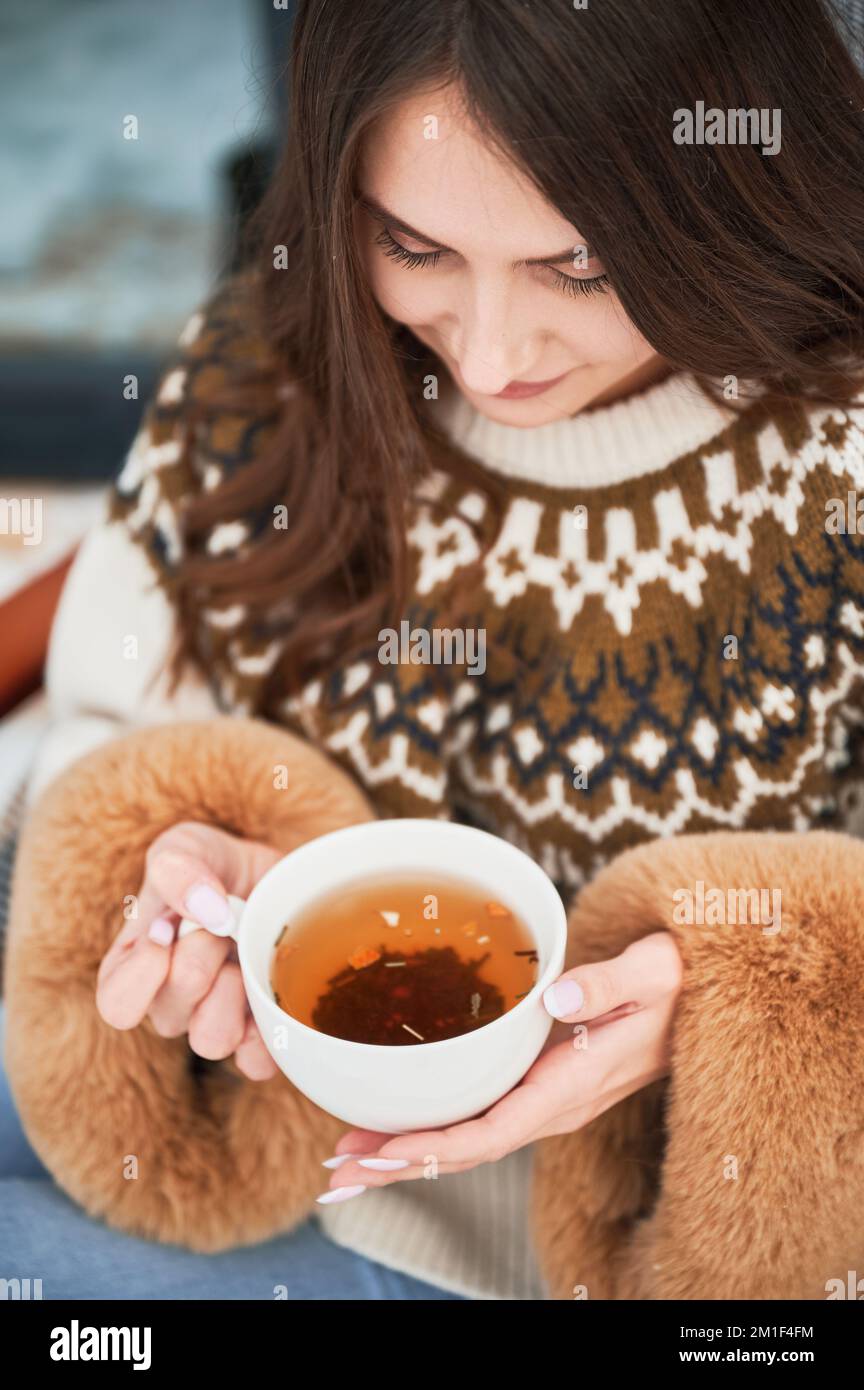 Primo piano di una felice femmina turista che tiene una tazza di tè, godendosi in un nuovo cottage in inverno. Giovane donna che riposa sulla terrazza della moderna casa fienile in montagna. Foto Stock