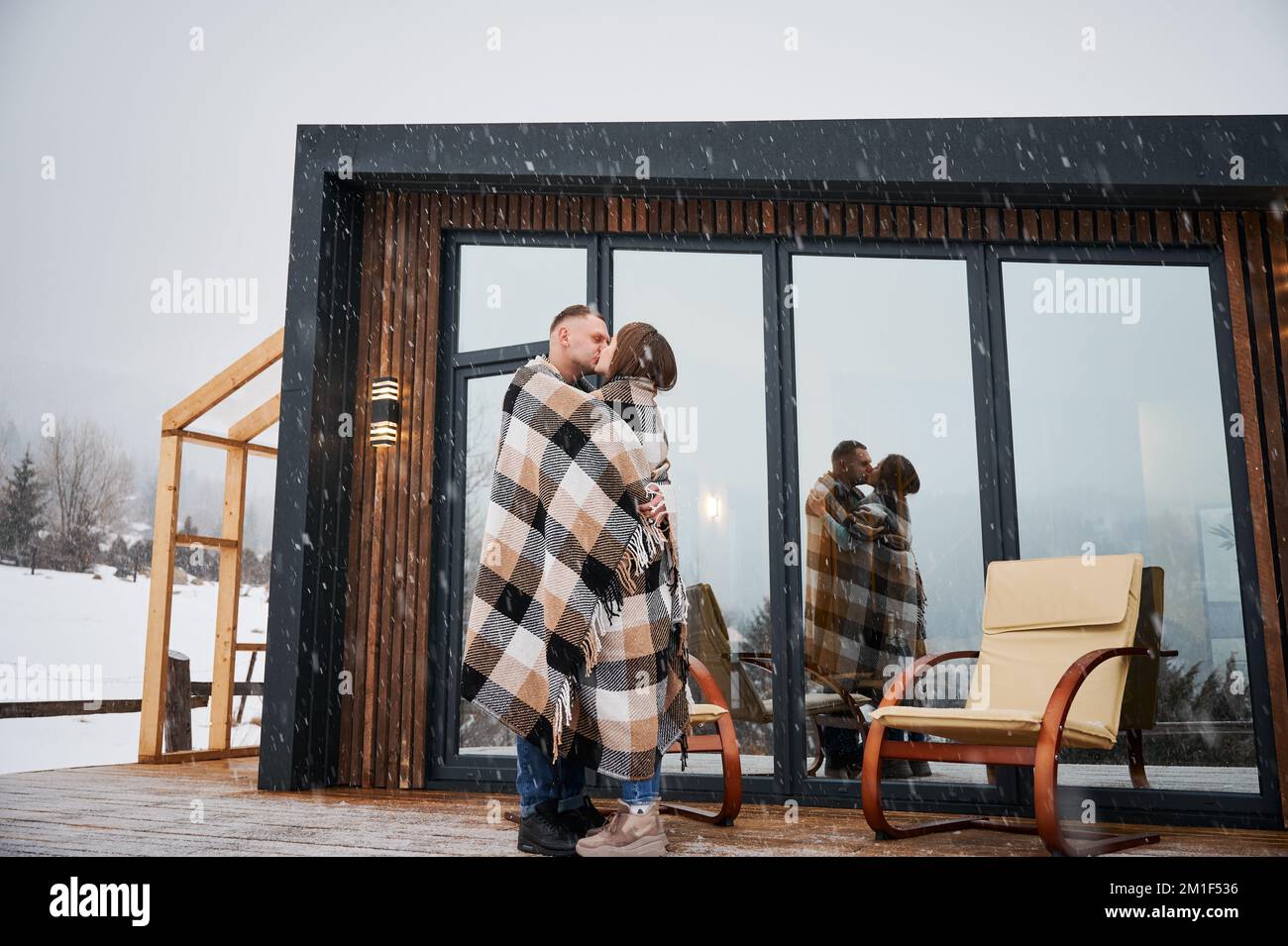 Uomo e donna avvolti in coperta baciandosi all'aperto sotto la neve d'inverno. Felice coppia che condivide il momento romantico mentre si trova vicino alle sedie fuori casa scandinava barnhouse con finestre panoramiche. Foto Stock