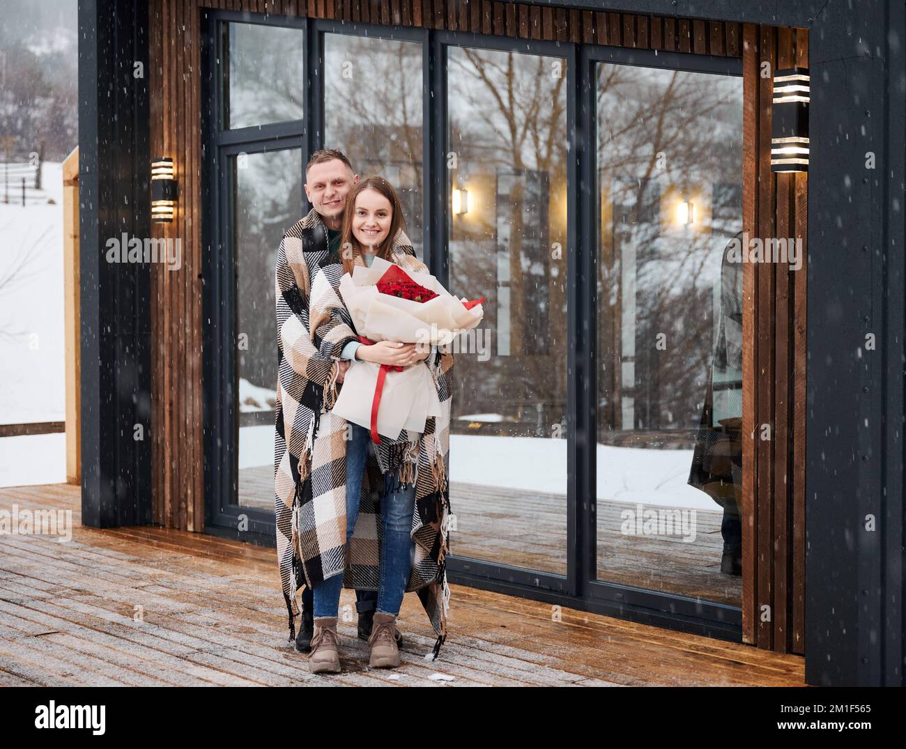 Tutta la lunghezza di coppia felice in piedi amore fuori casa scandinava barnhouse sotto la bella neve. Uomo coperto di coperta abbracciante ragazza e sorridente mentre donna in possesso bouquet di rose. Foto Stock