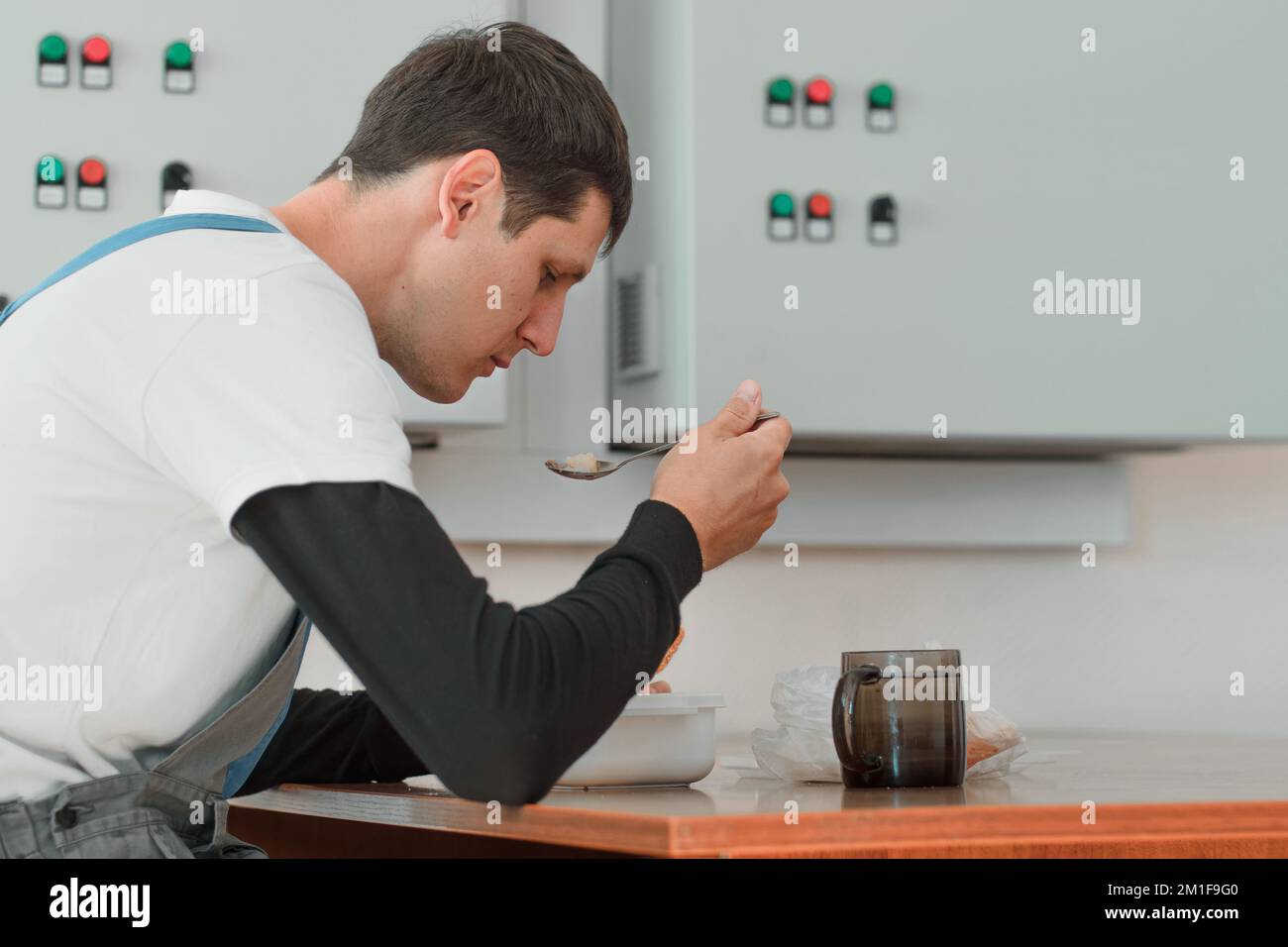 Pausa pranzo. Mangiare sul posto di lavoro durante il lavoro. L'uomo caucasico in tuta da lavoro siede al tavolo nella sala di produzione e mangia dal contenitore. Il lavoratore ha pranzo in sala pausa. Foto Stock