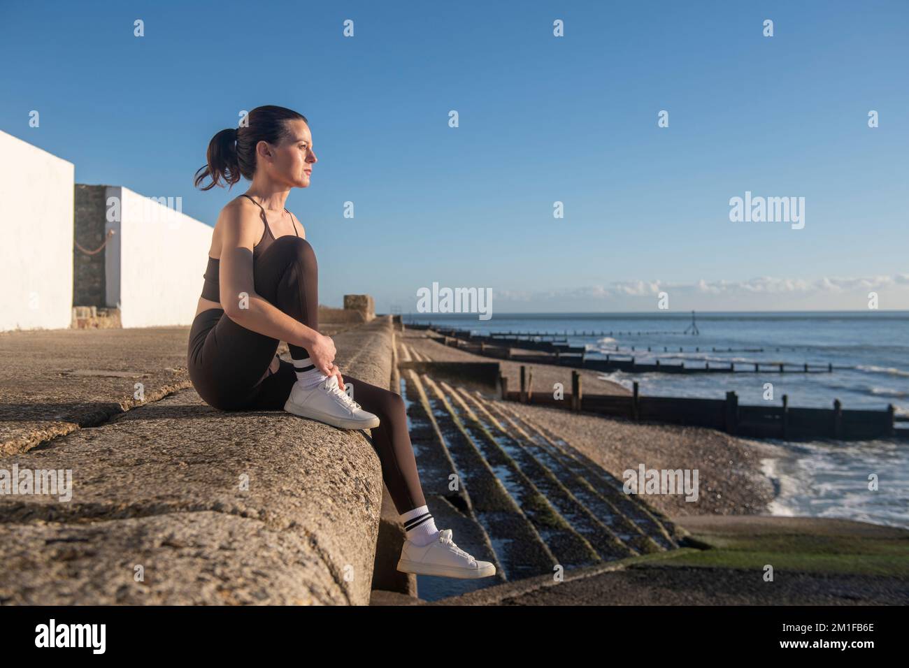 Donna sportiva seduta su un muro in riva al mare, dopo essersi allenata all'aperto. Godendo il sole e la vista. Foto Stock