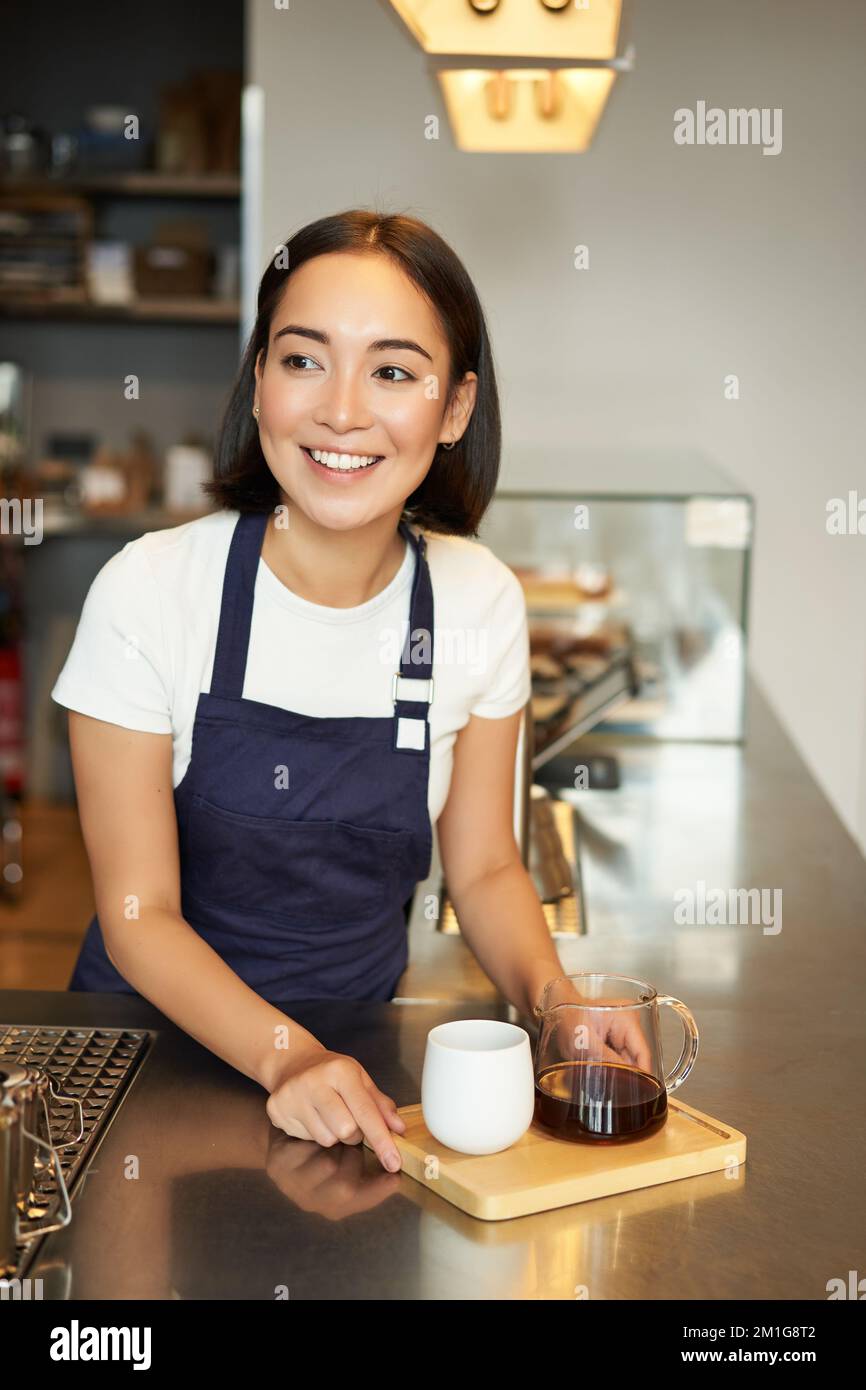 Immagine verticale di una ragazza sorridente barista che serve caffè, prepara una birra a lotti, filtra per i clienti nel bar, indossa un grembiule blu dietro il bancone Foto Stock