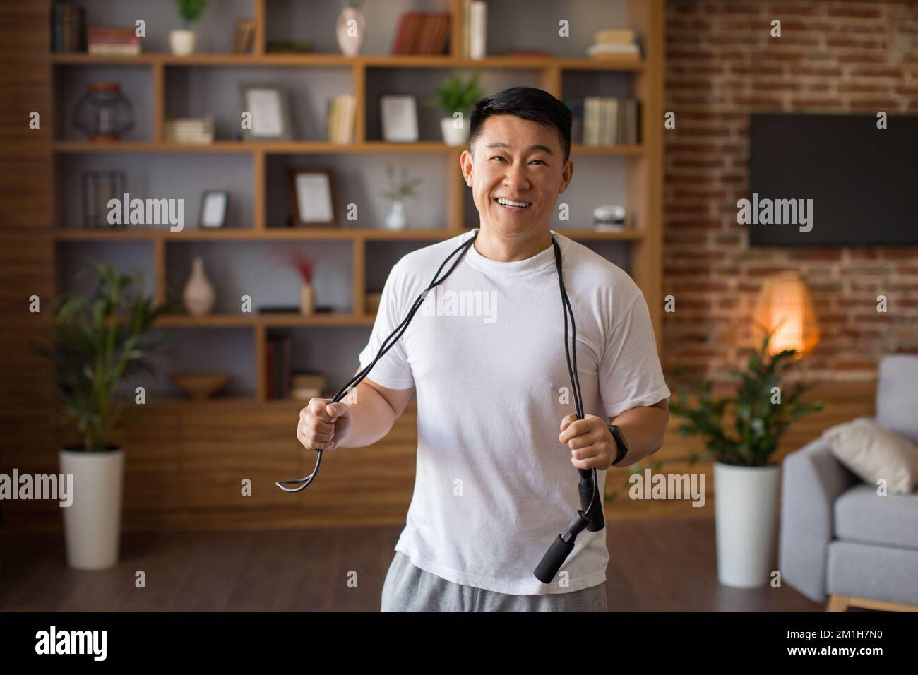 L'eccitato uomo asiatico di mezza età in piedi con la corda saltante appendere intorno al collo, pronto ad allenarsi a casa in salotto Foto Stock