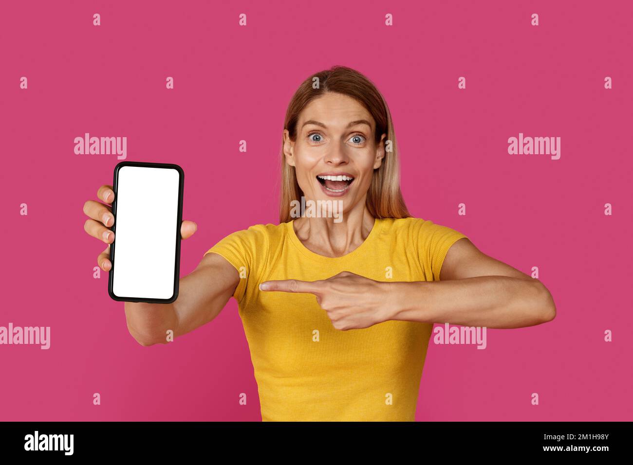 Felice sorpresa ispirata donna matura caucasica bionda in t-shirt gialla con bocca aperta punta dito sullo smartphone Foto Stock