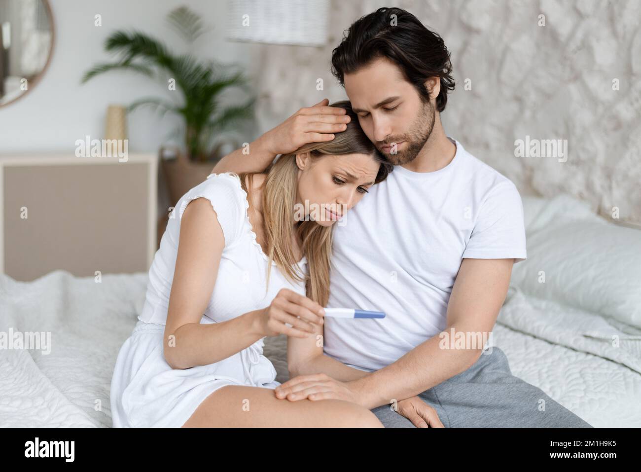 Problema di infertilità. Coppia caucasica seduta a letto con esito negativo del test di gravidanza Foto Stock