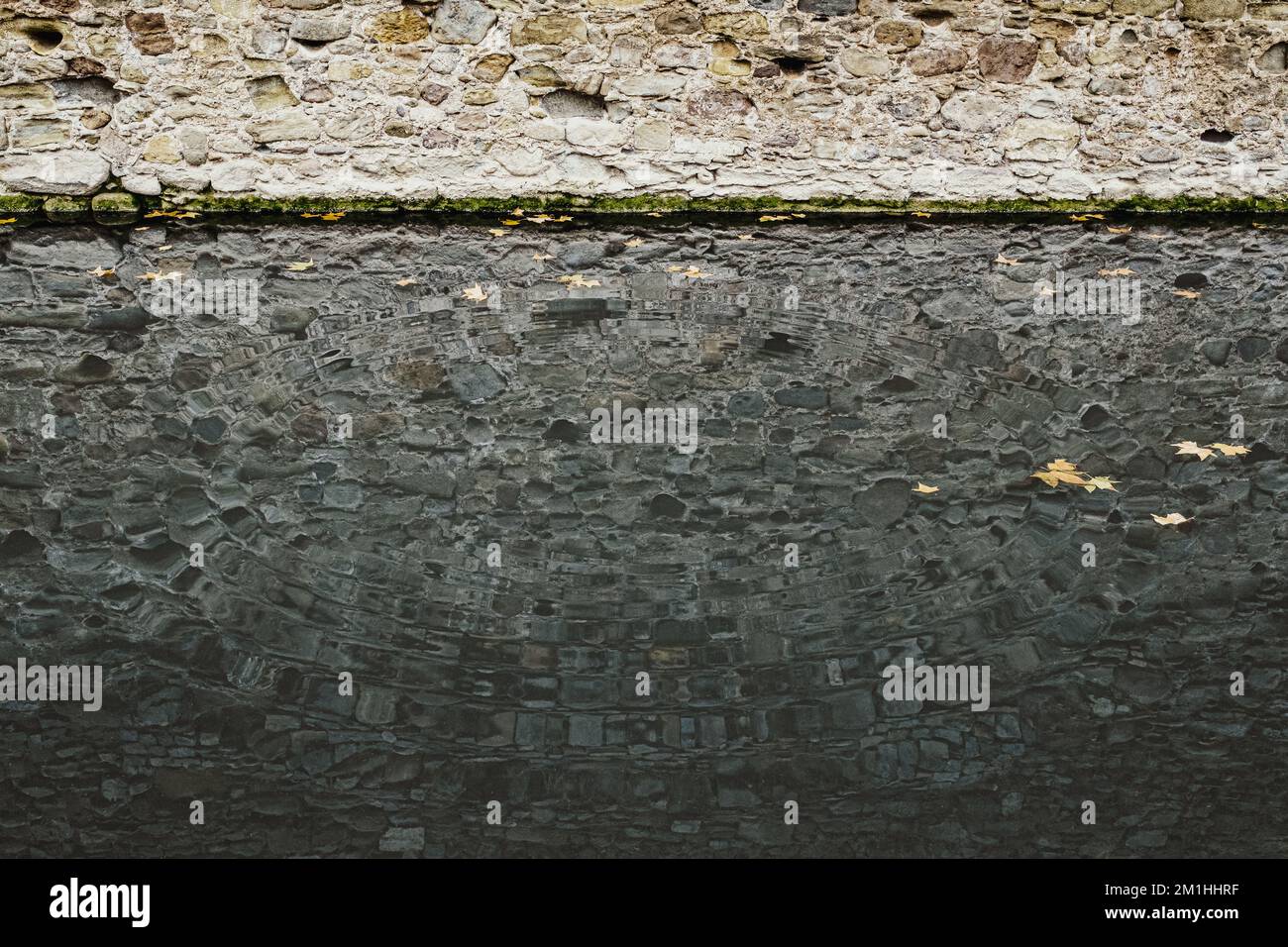 Andamento ad onda concentrica nell'acqua del torrente e riflesso dell'antica parete di pietra lungo la riva. Foto Stock