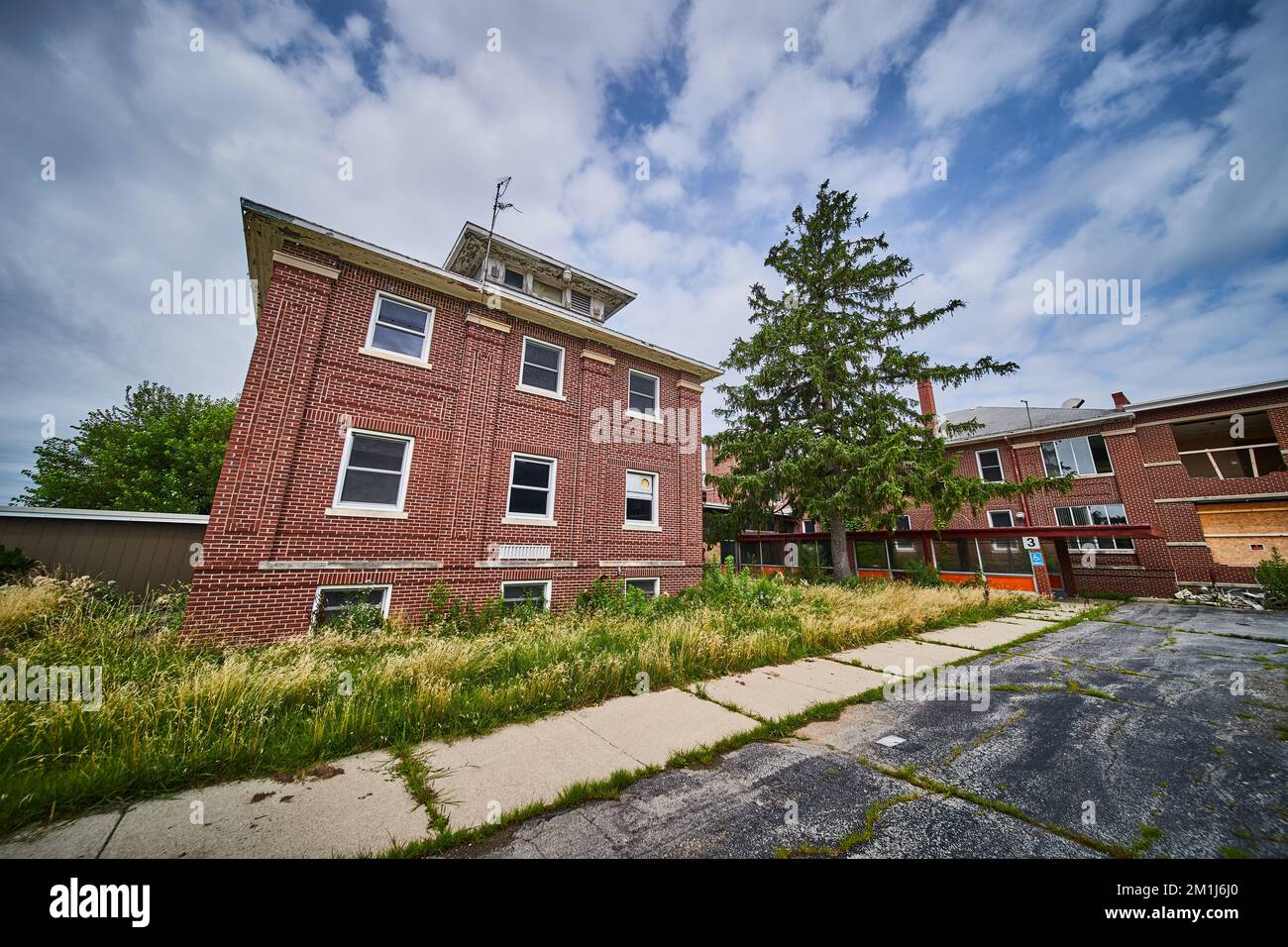 Edificio a tre piani abbandonato in mattoni dal parcheggio esterno con campi troppo grandi Foto Stock