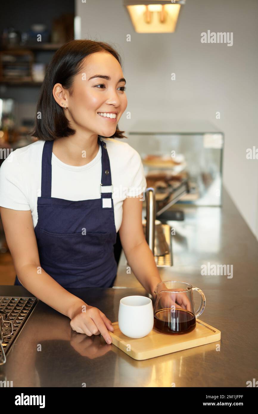 Immagine verticale di una ragazza sorridente barista che serve caffè, prepara una birra a lotti, filtra per i clienti nel bar, indossa un grembiule blu dietro il bancone Foto Stock