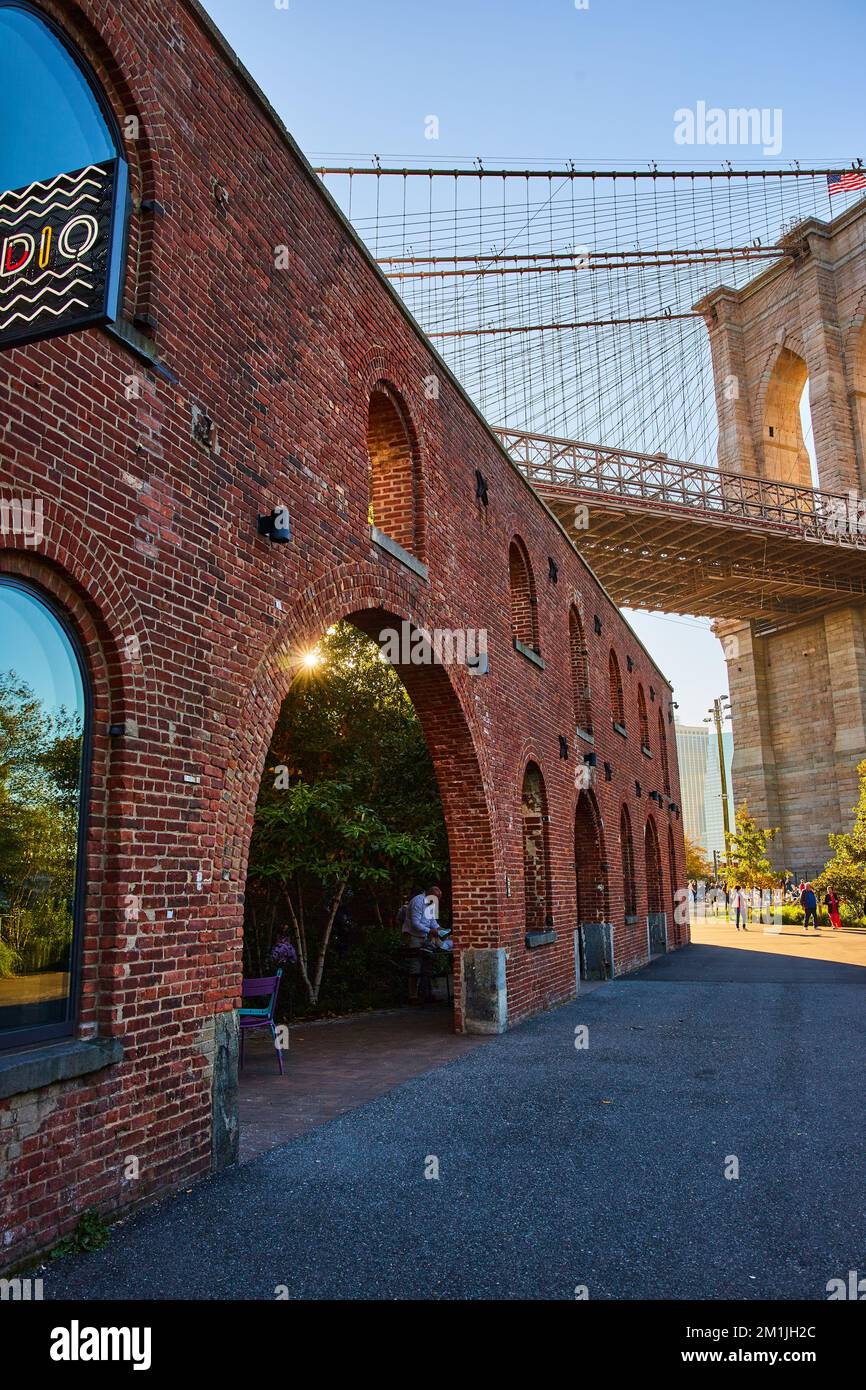 Edificio in mattoni con archi e sole dorato che sorpassa accanto allo splendido e iconico Ponte di Brooklyn a New York Foto Stock