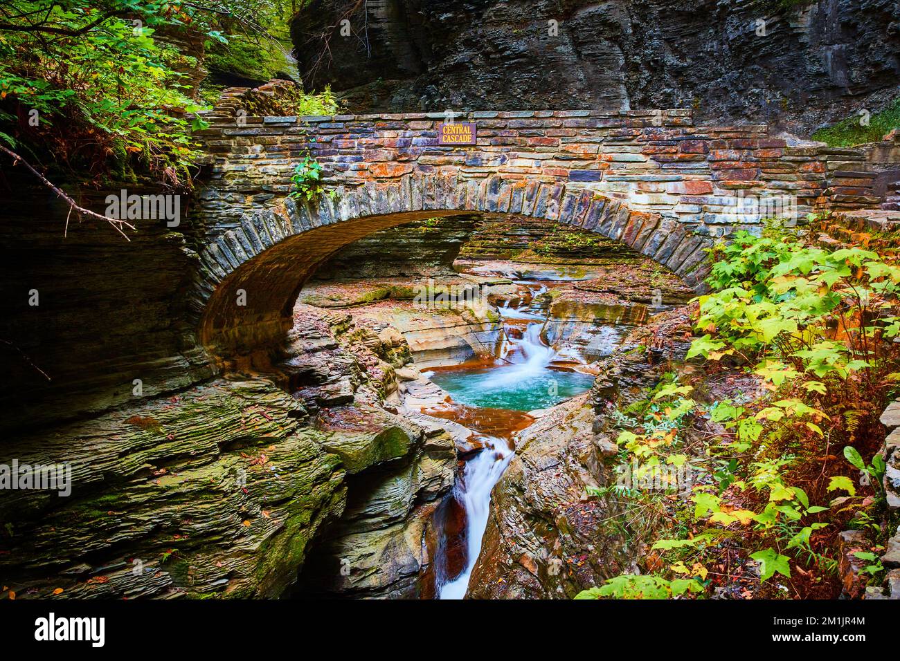 Bellissimo ponte a piedi arco di pietra sopra gola e fiume con acque blu e cascate durante la caduta Foto Stock