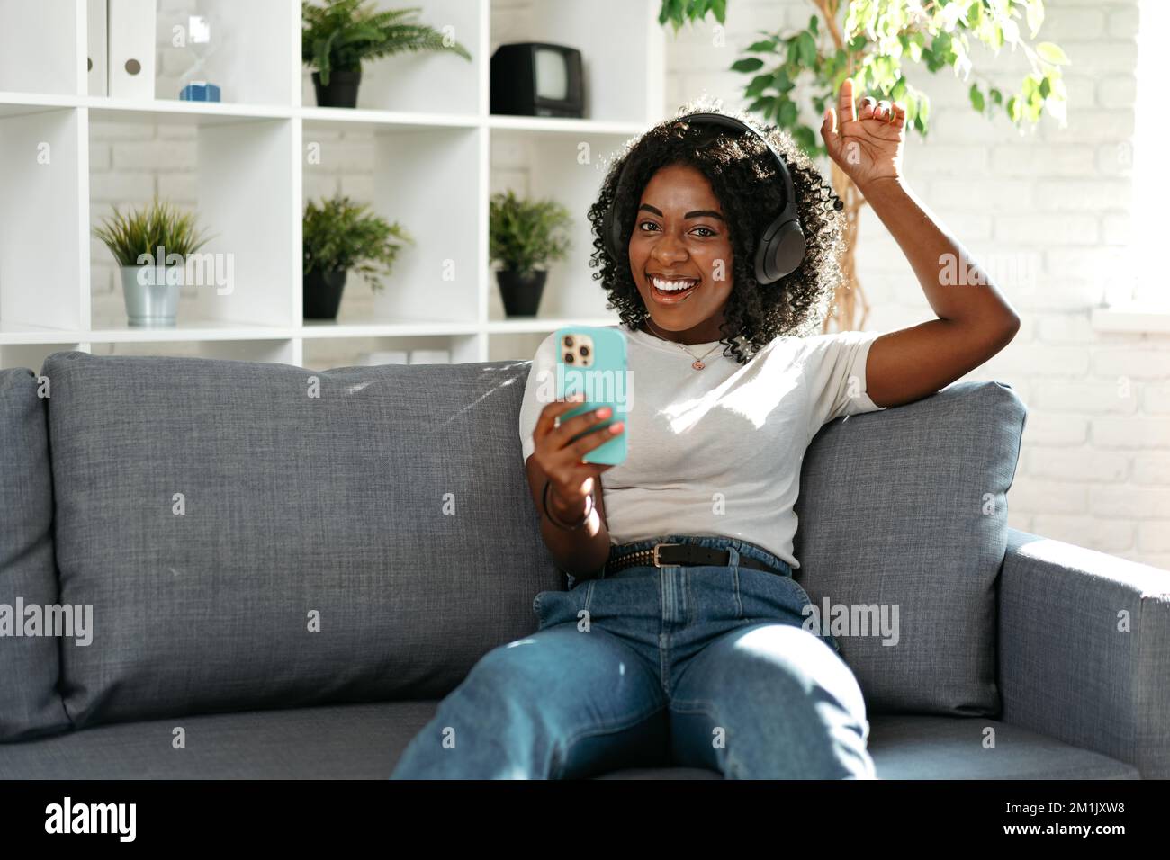 Donna africana sorridente seduta sul divano a casa e con lo smartphone Foto Stock