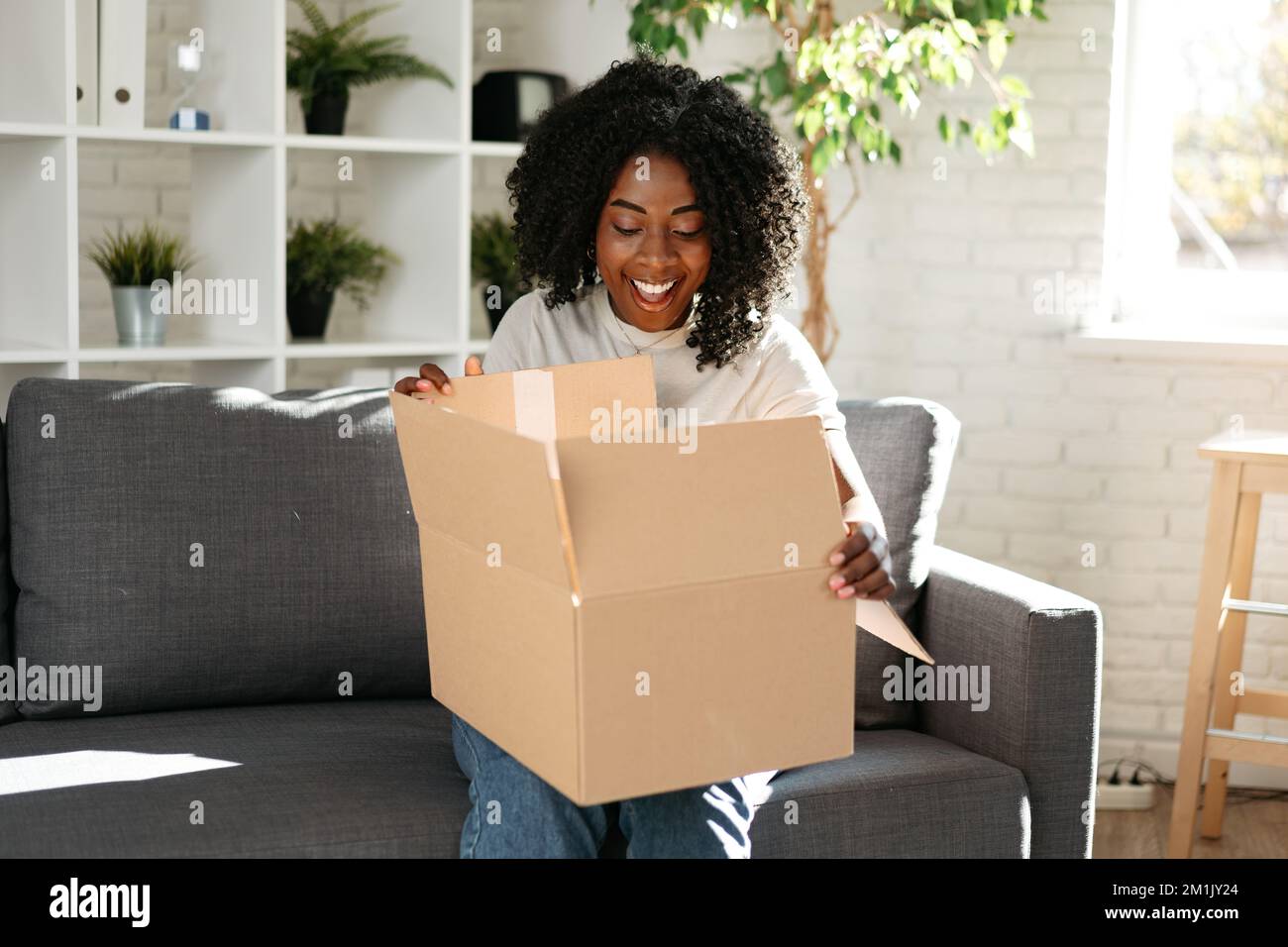 Giovane donna africana sedersi sul divano a casa disimballando pacco scatola di cartone con l'acquisto online Foto Stock