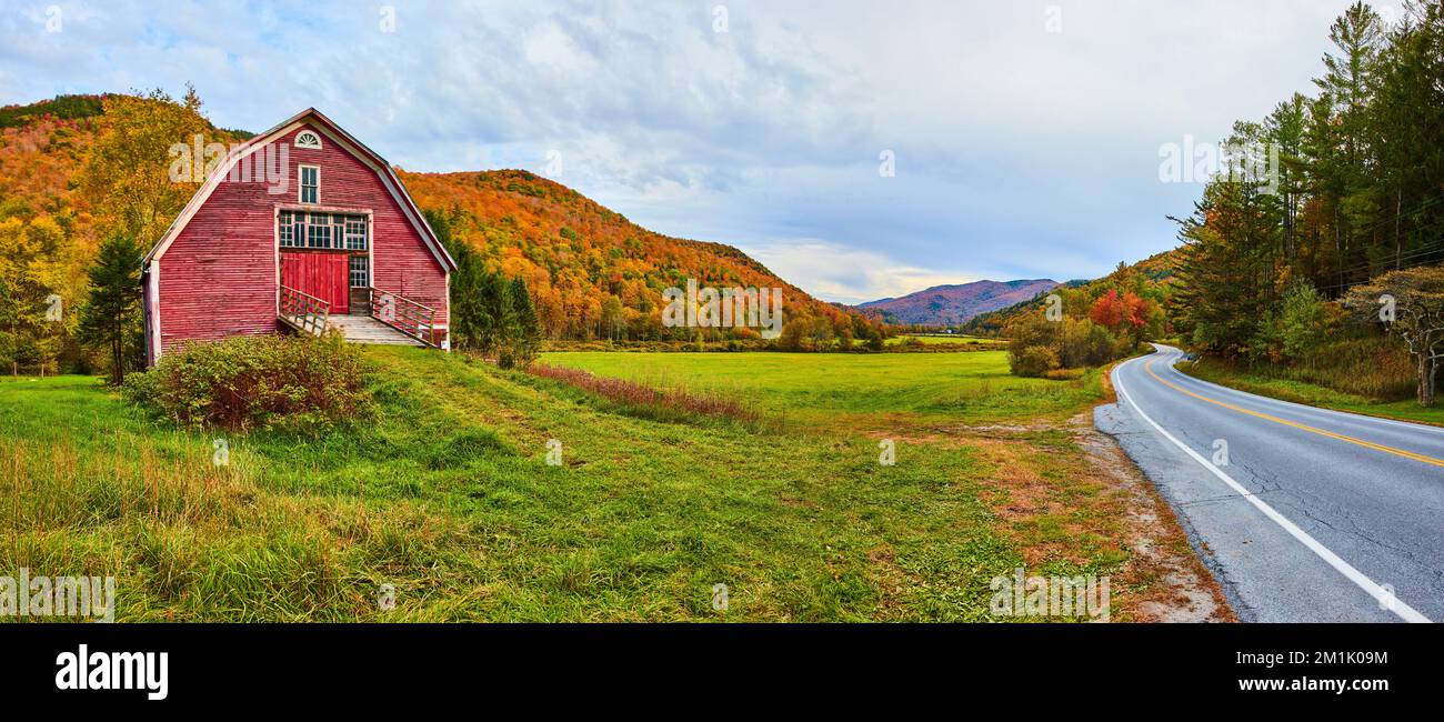 Vista panoramica sulla strada nel Vermont del fienile rosso in campi erbosi circondato da montagne fogliame caduta Foto Stock