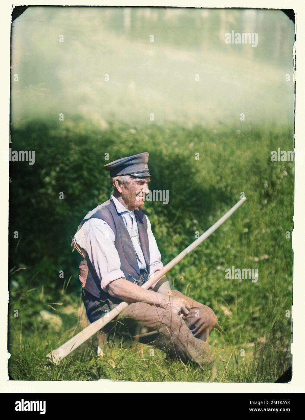Fotografia colorata vintage di un vecchio seduto in un campo in un giorno di sole estati indossando un vecchio cappuccio miitario che tiene un remo. circa 1900 Foto Stock