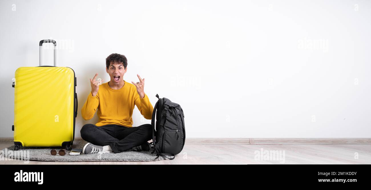 Giovane e bel viaggiatore con valigia che indossa un maglione seduto su sfondo bianco urlando con pazza espressione facendo simbolo rock con le mani in su Foto Stock