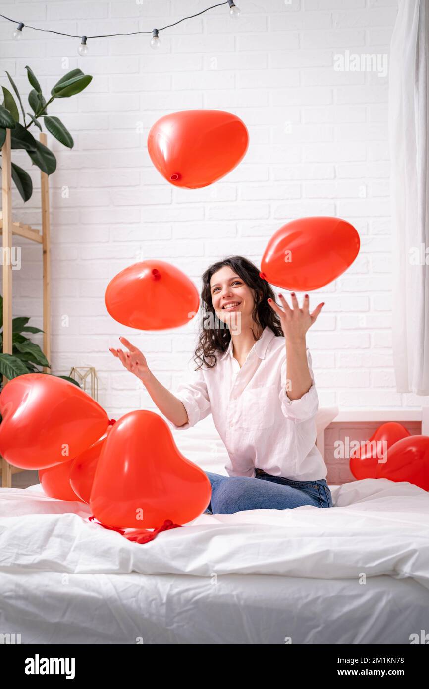 Brunette ragazza europea gioisce giocando a letto con palloncini rossi a forma di cuore. Omaggio sorpresa dell'amore del mattino il giorno delle valentine Foto Stock