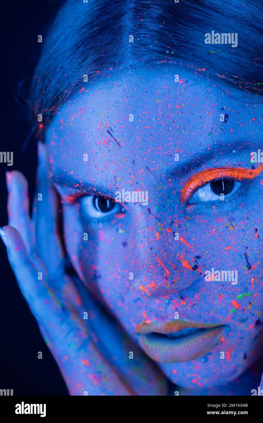 primo piano ritratto di donna con il trucco neon e vernice colorata sul viso guardando la fotocamera isolato su blu scuro, immagine stock Foto Stock