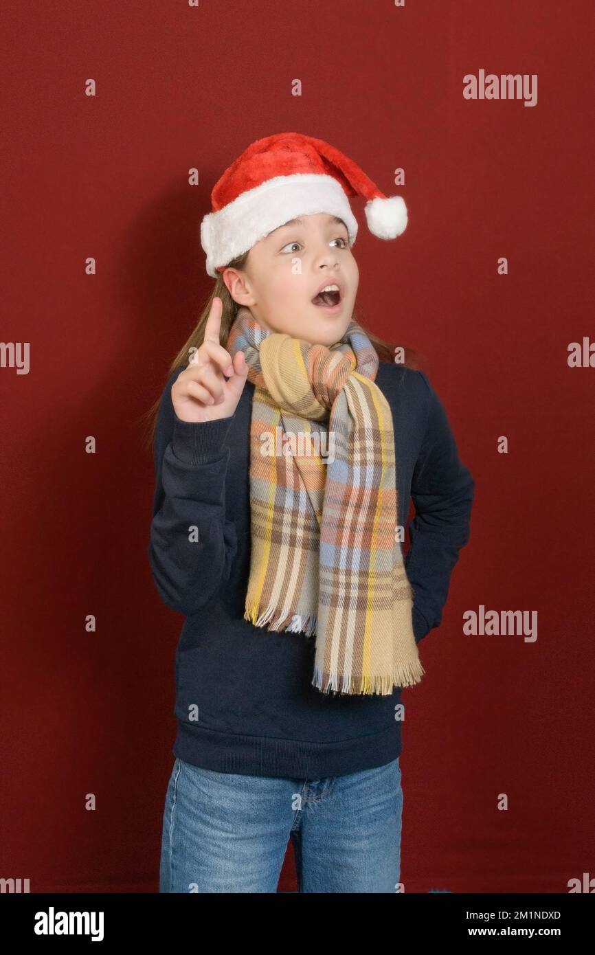 Nuova idea per natale. Ragazza adolescente in cappello di santa e sciarpa plaid, indice dito in su. Sfondo scuro bordeaux Foto Stock