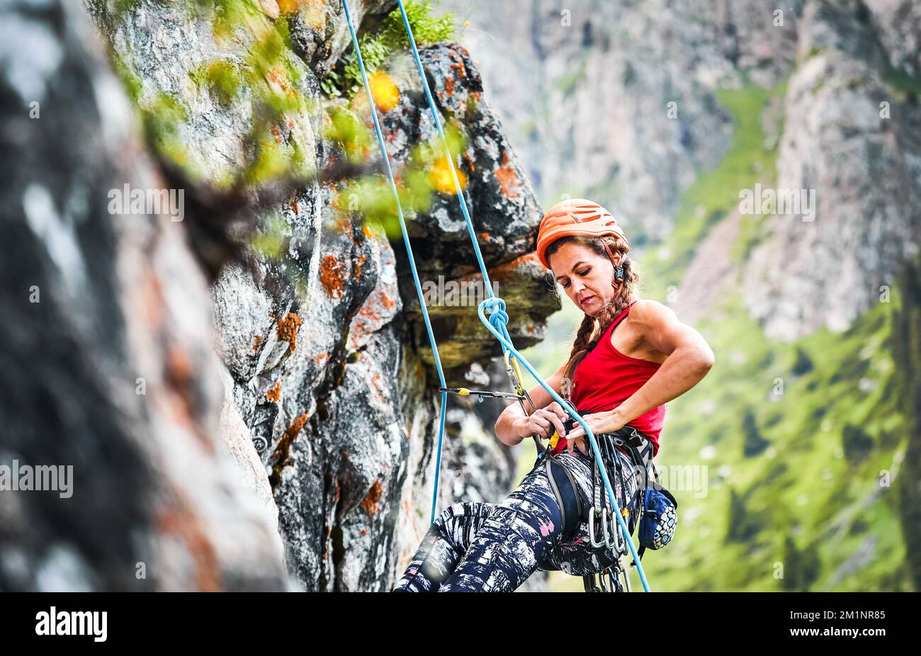 Ritratto di Fit donna forte in pantaloni rossi e casco arrampicata sulla roccia alta verticale con corda in primo piano sulle montagne Foto Stock