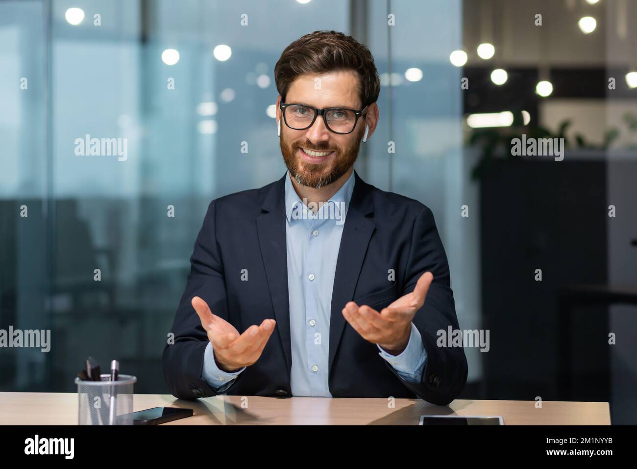 Ritratto di un giovane e simpatico uomo seduto in ufficio al tavolo indossando le cuffie e parlando con la fotocamera. Incontro d'affari, supporto al tutor, colloquio di lavoro, consulenza Foto Stock
