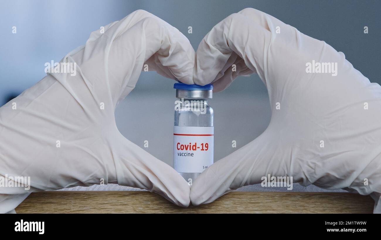 Primo piano della mano femminile in lattice bianco guanti medici facendo forma del cuore mostra segno di amore speranza affidabilità approvazione vicino bottiglia di vaccino dose da Foto Stock