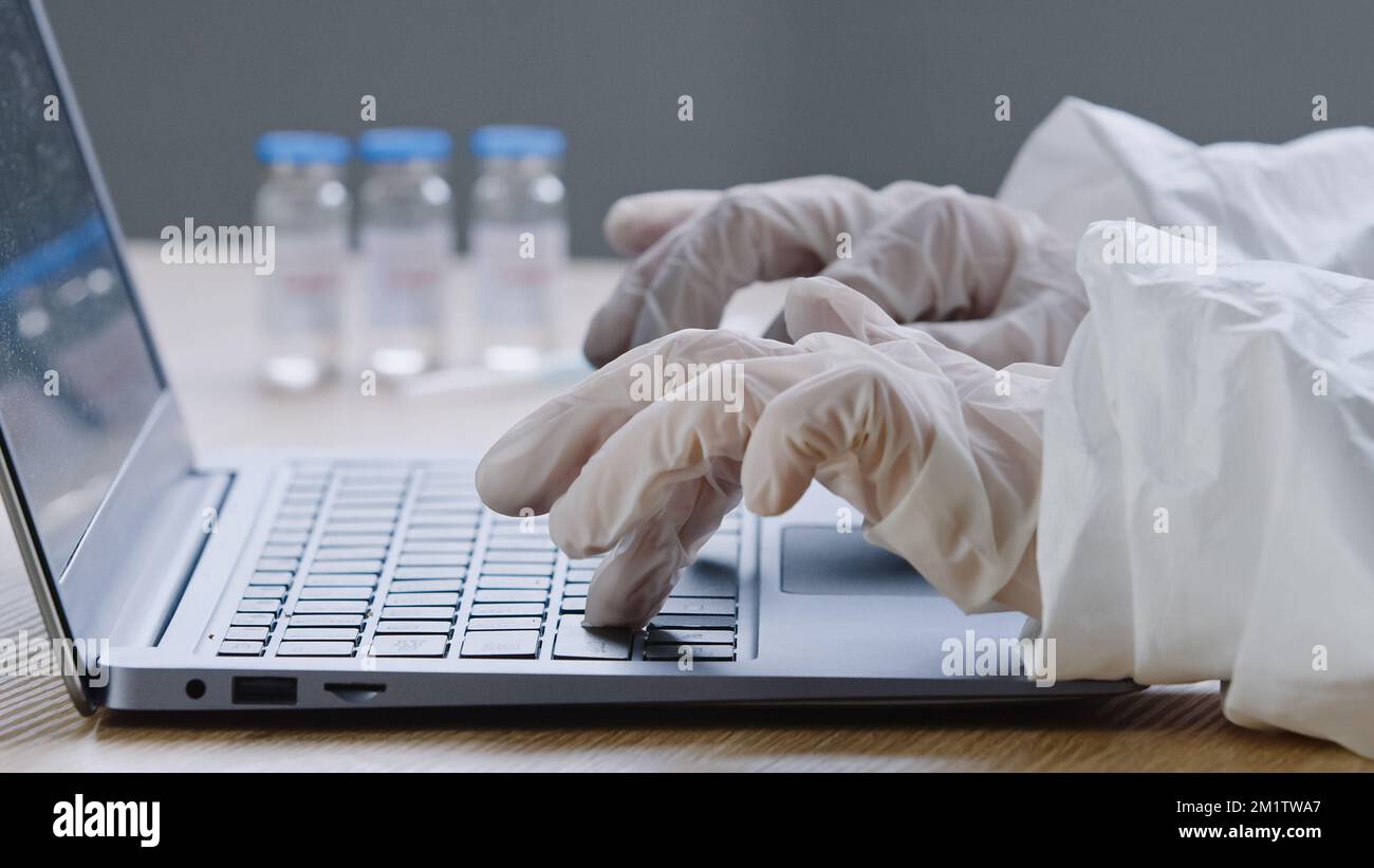 Primo piano del vaccino covid-19 si trova su un tavolo di legno in clinica medico irriconoscibile scienziato in guanti di lattice digitando su laptop marche Foto Stock