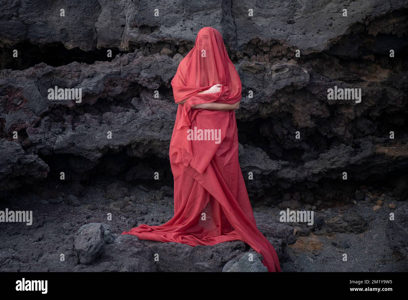 Giovane donna coperta da un lungo velo rosso da dovette ai piedi guardando in su con gli occhi chiusi mentre si trova di fronte ad una parete rocciosa Foto Stock