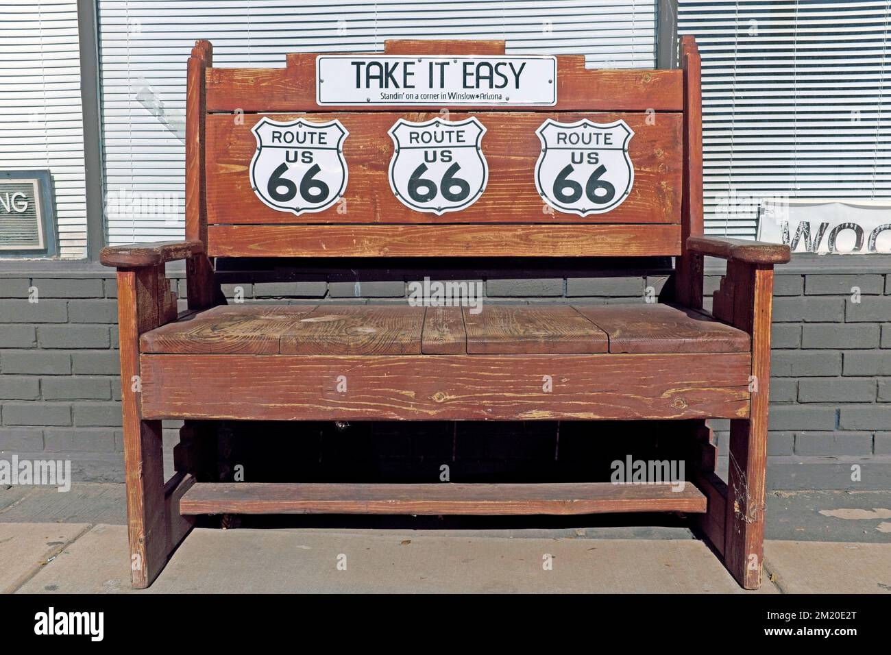 Una panca di parco in legno si trova sulla Route 66 nel centro di Winslow, Arizona, USA, con tre segnali stradali per la Route 66 e un cartello con la scritta "Take IT Easy". Foto Stock
