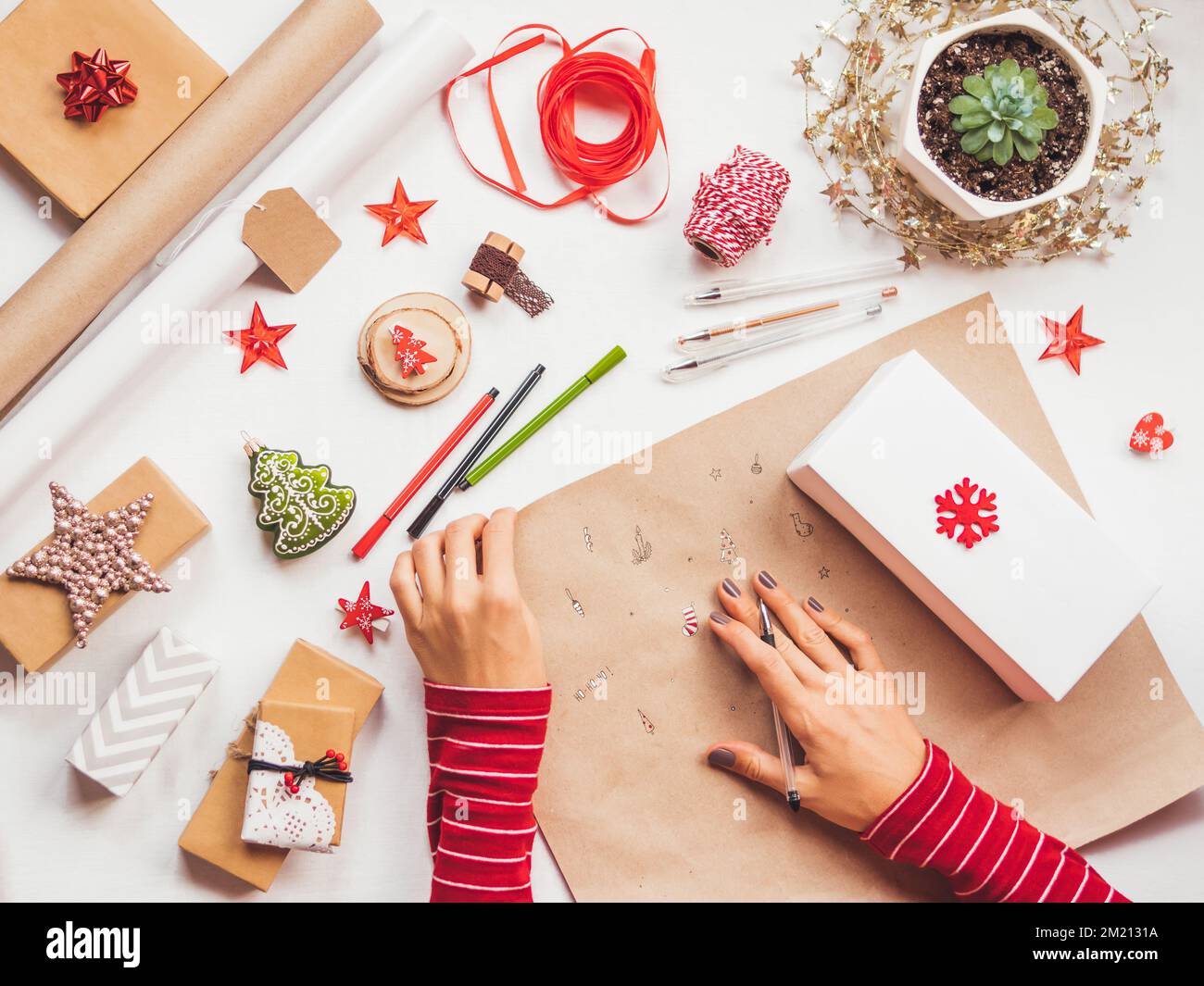 Vista dall'alto sul tavolo con decorazioni natalizie. Donna disegna i simboli di Capodanno su carta artigianale e avvolge regali. Giacitura piatta. Foto Stock