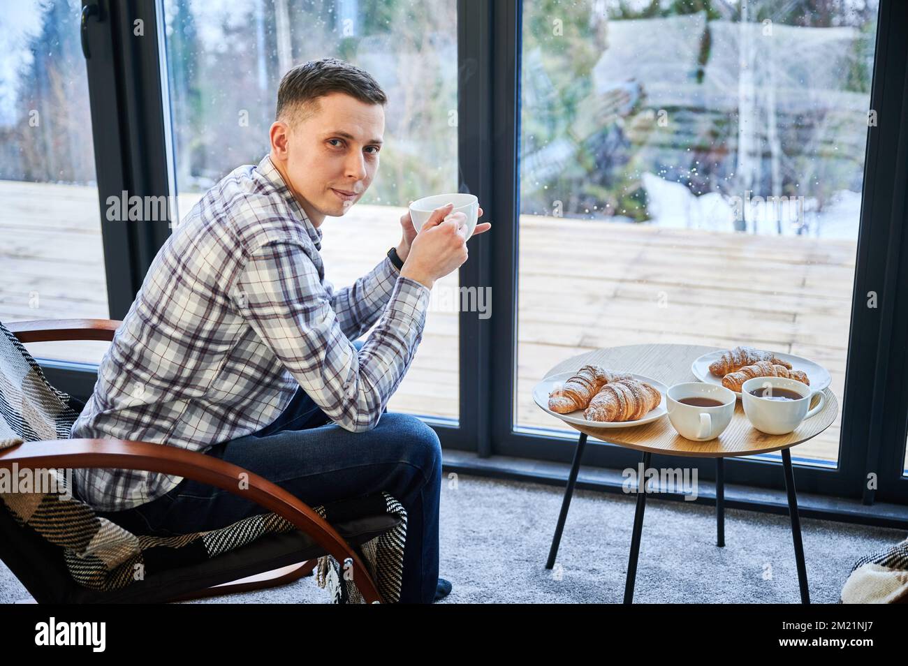 Giovane uomo che si gode week-end all'interno di una caserma contemporanea. Ritratto di turista maschile seduto su sedia, tenendo una tazza di tè in cottage nuovo. Foto Stock