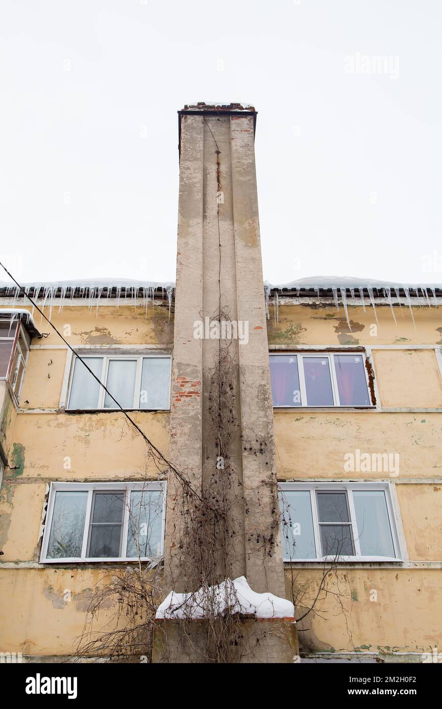Fumo, tubo di scarico in pietra lungo il muro della vecchia casa. Sul tetto sono presenti neve e ghiaccio, sullo sfondo di un cielo grigio. Nuvoloso, freddo giorno d'inverno, luce soffusa. Foto Stock
