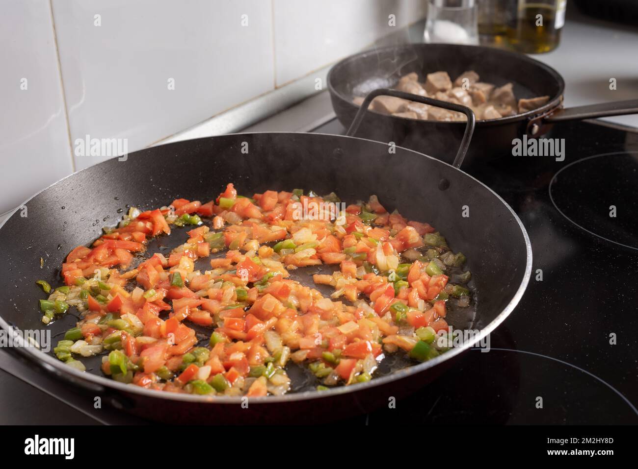 Padella per paelle con verdure in camicia, sullo sfondo un'altra padella con carne sul piano cottura in casa. Foto Stock